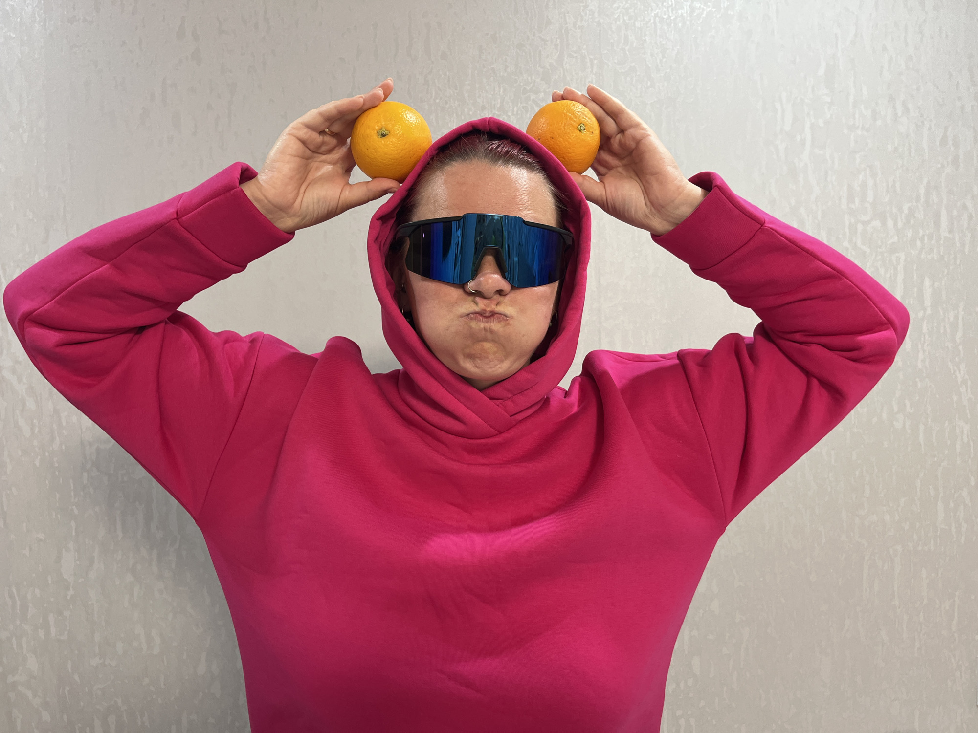 A person with pink hair is indoors posing for a photo in front of a light gray patterned wall. The person is wearing a bright pink hoodie, the hood pulled up. Large, reflective blue sunglasses cover their eyes. They are holding an orange in each hand on top of their head, creating the appearance of mouse ears. Their face is puckered in a silly expression. The exact location in Chaikovsky, Russia is indiscernible from the image.