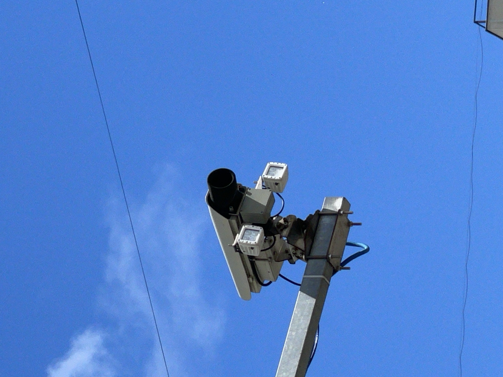 A speed camera is mounted on a metal pole against a clear blue sky with thin wisps of clouds. The camera system consists of a primary unit with a large, dark cylindrical lens, accompanied by two smaller white boxes, likely sensors or additional cameras. The equipment is attached to the pole with metal brackets and bolts, with a blue wire visible. Another thin black wire runs diagonally across the sky from the top left to the bottom right. In the far upper right corner, a sliver of a white structure, possibly a building or another pole with an attachment, is visible. The scene suggests it is daytime, likely sunny due to the clear sky. No people or specific Moscow landmarks are identifiable.