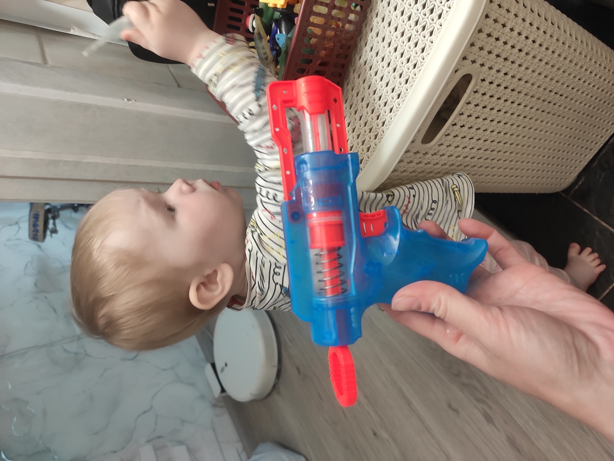 A child lies on a tiled floor, positioned next to a white storage container and a brown wicker basket filled with toys. The child, with fair hair, is wearing a striped shirt. An adult hand holds a blue and red toy gun, pointing it towards the child. Part of an adult foot is visible in the background. The location appears to be indoors, possibly a bathroom or a utility area. There is insufficient information to confirm that this image was taken in the city of Sterlitamak, Russia.