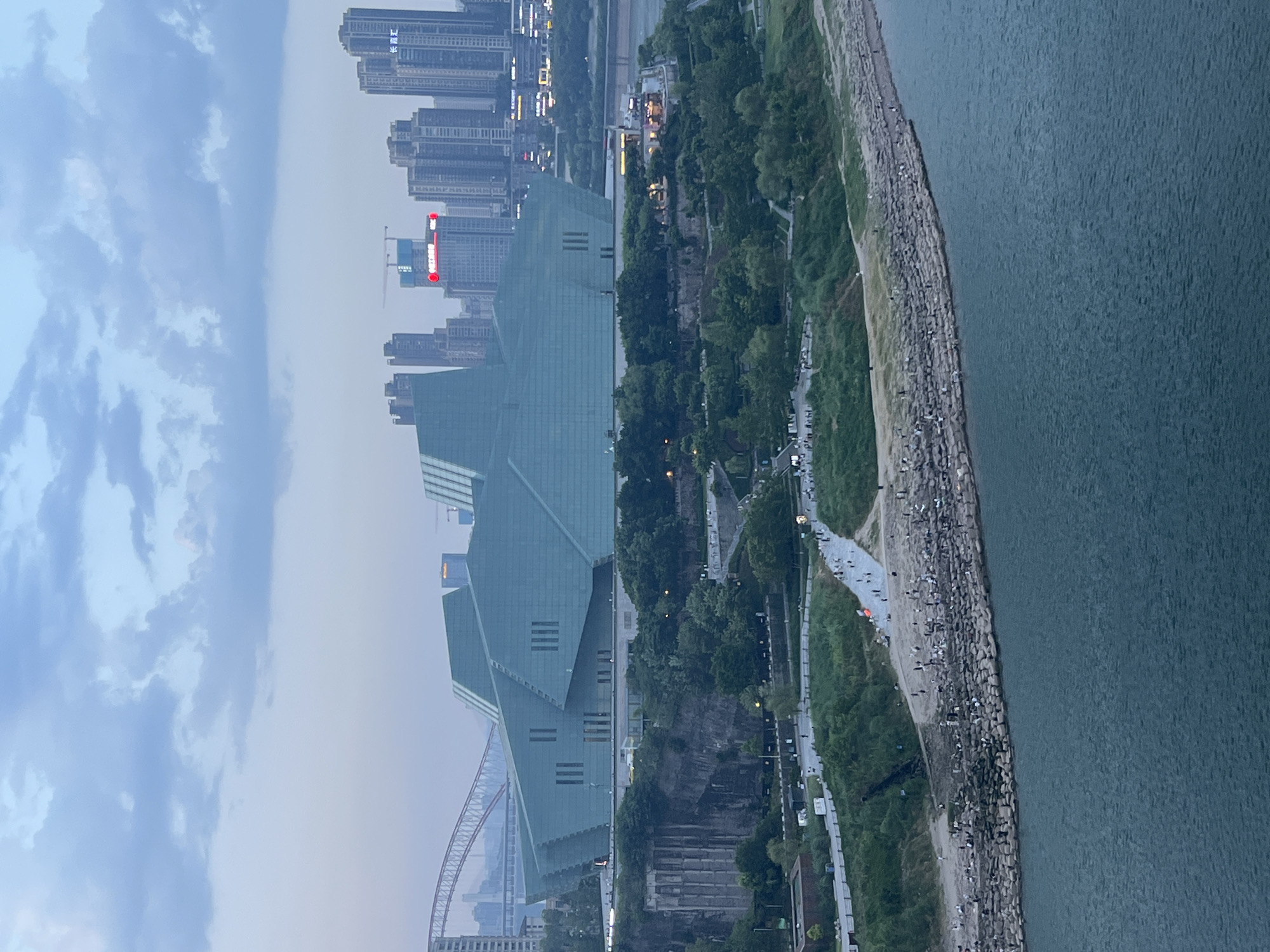The image shows a wide view of a waterfront with a large, modern building with a greenish-blue facade prominently in the foreground. The building has a geometric, multi-faceted design. To the left of the building, there is a steep, rocky cliff face. In front of the building and extending along the waterfront, there are areas of greenery, pathways, and a rocky embankment where numerous people are gathered. The water in the foreground is a dark blue-green. In the background, a cityscape with tall buildings and a bridge can be seen under a cloudy sky. The location is identified as Jiangbeicheng Street, China.