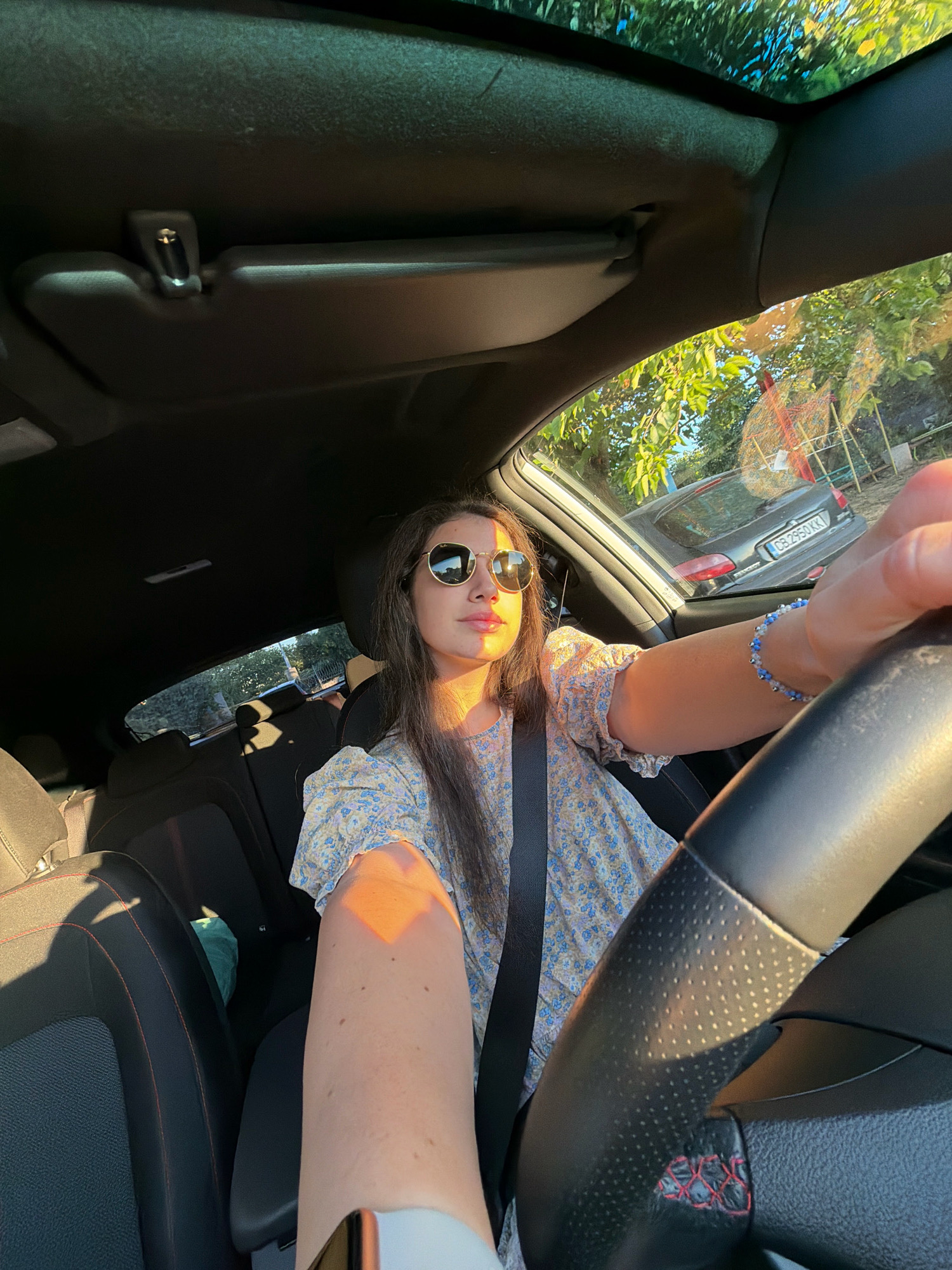 The image depicts a young woman driving a car. She is wearing sunglasses and a floral-patterned dress. The interior of the car is visible, including the steering wheel, seats, and a sun visor. The exterior of the car shows trees and another vehicle with a license plate that reads "CB 2950 KK". This license plate format is consistent with vehicles registered in Bulgaria, and specifically with the Sofia region. The lighting suggests it is either sunrise or sunset, casting a warm glow on the scene. The image appears to be a selfie taken by the driver.