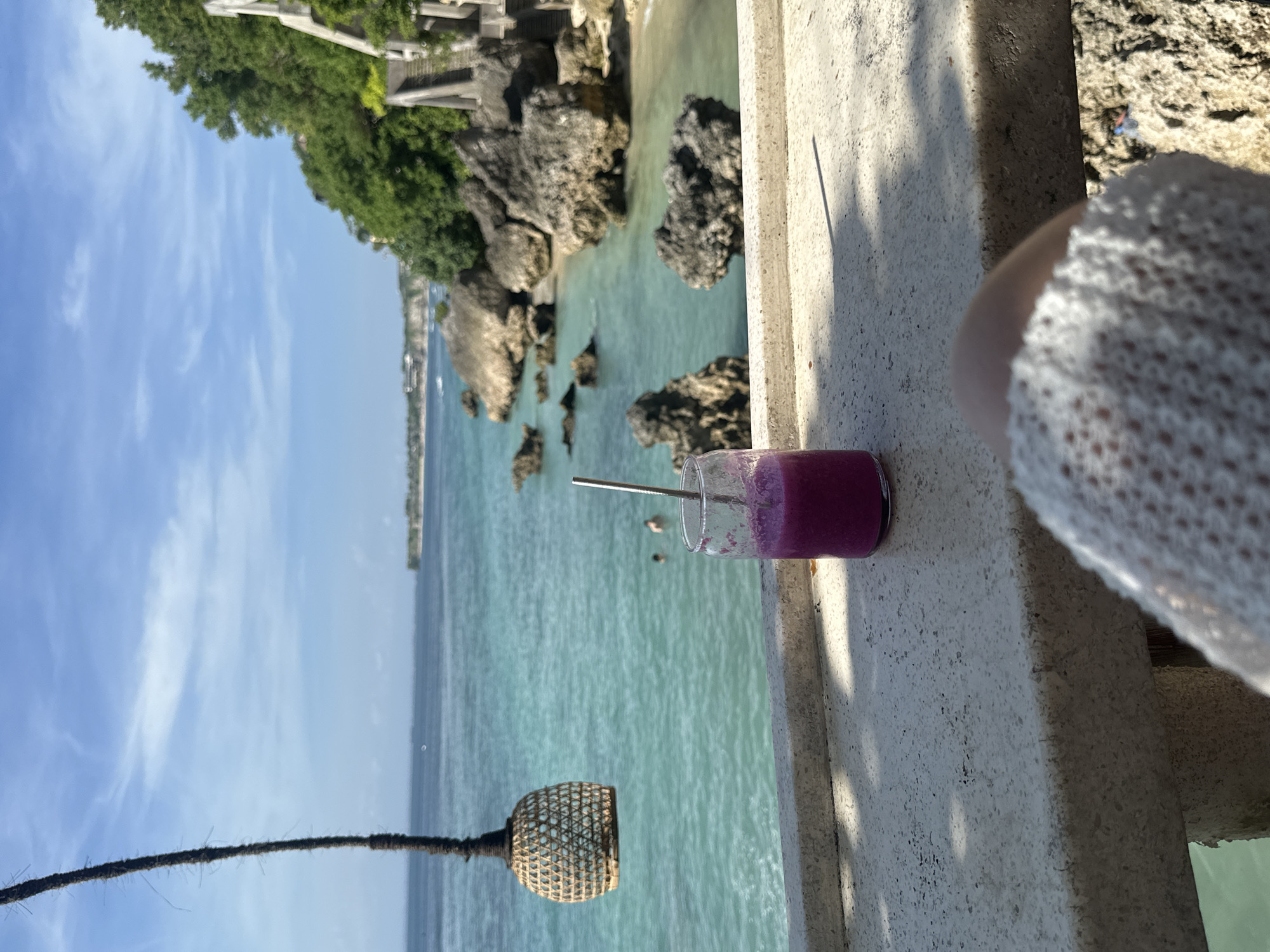 The image shows a purple drink in a glass with a metal straw, placed on a concrete surface with a beach and ocean view in the background. The ocean is turquoise with visible rocks and a few distant figures in the water. A lamp hangs below the drink. Green vegetation and rock formations are visible along the coastline. The image appears to be taken in Kuta Selatan, Indonesia. A person wearing a white garment is partially visible in the foreground.