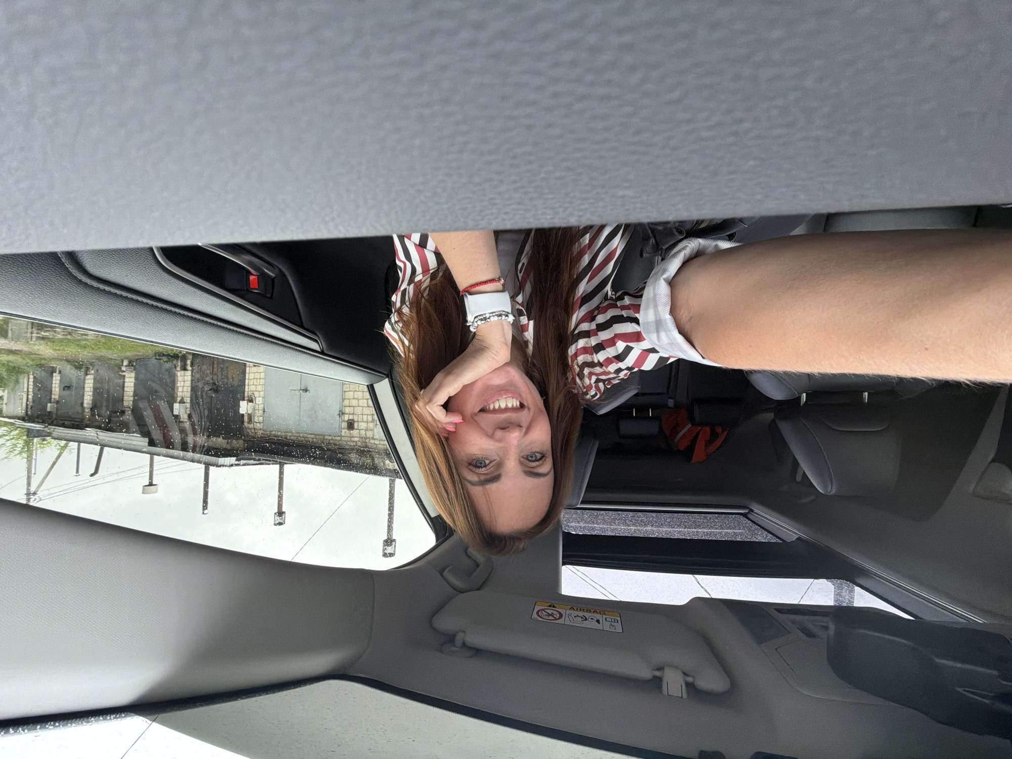 A woman with shoulder-length brown hair and blue eyes smiles at the camera from the driver's seat of a car. She is wearing a red, white, and black striped shirt and has her hand resting on her chin. The car has a sunroof open, and it appears to be raining outside, as evidenced by water droplets on the windshield and the overcast sky. In the background, a row of garages with metal doors can be seen. The location is identified as Birobidzhan, Russia, a city in the Jewish Autonomous Oblast known for its agricultural and industrial sectors. The woman is holding a smartphone in her left hand, taking a selfie. The interior of the car is visible, including the dashboard and part of the passenger seat. An orange safety vest is visible on the passenger seat.
