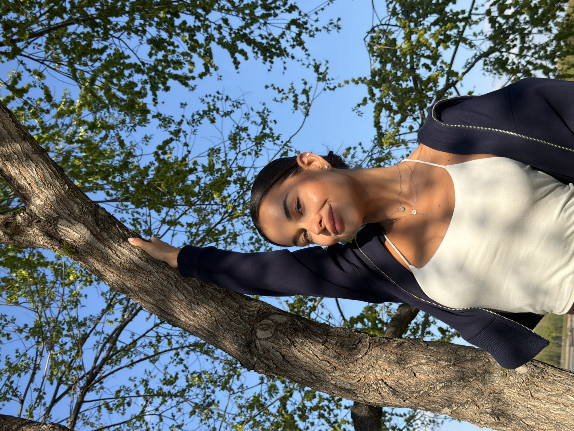 A young woman with dark hair pulled back, wearing a white tank top and navy blue zip-up jacket, is leaning against a thick tree branch in a sunny outdoor setting. The sky is blue with leafy trees filling the background. The photograph appears to be taken at a low angle, looking up towards the woman and the sky. The location is consistent with an outdoor park in Krasnoyarsk, Russia given the type of trees, blue sky and geographical location of the subject.