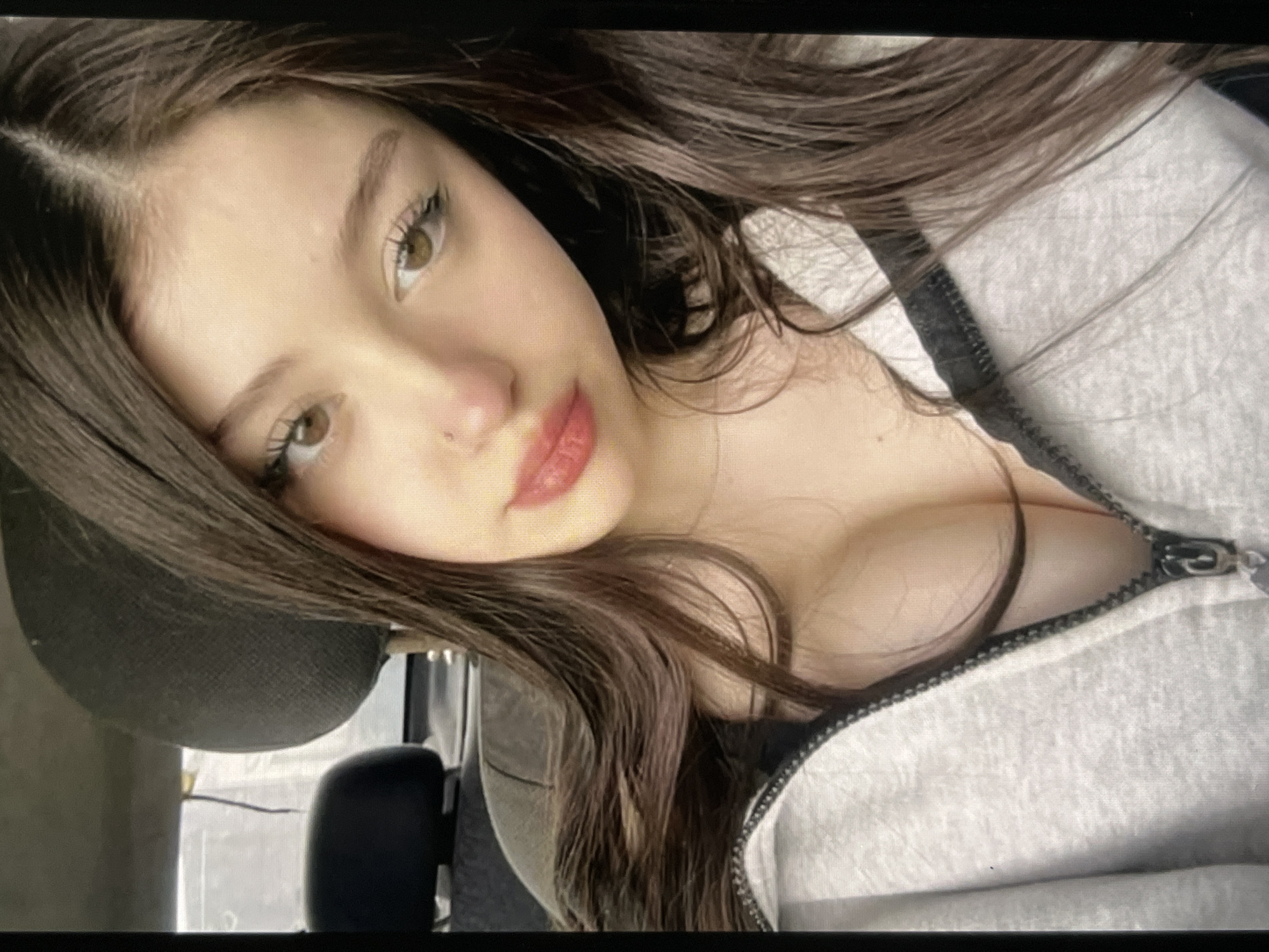 A young woman with long, dark brown hair is pictured from the chest up, seated in what appears to be the driver's seat of a car. She is wearing a gray zippered hoodie, which is partially unzipped, revealing her chest. Her head is tilted slightly to the right, and her eyes are looking forward. She has light brown eyes, defined eyebrows, and noticeable eyelashes. Her lips are slightly parted and appear to be colored with a pinkish-red lipstick. A small nose ring is visible on her right nostril. The background is blurred, but suggests the interior of a vehicle, with car seats and a headrest visible. The lighting suggests it is daytime.