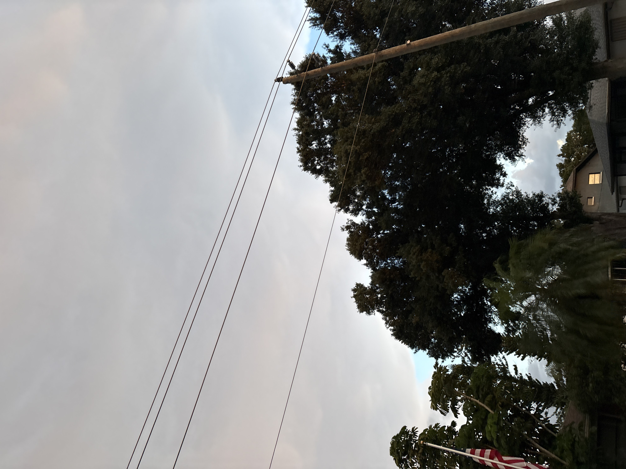 The image depicts a low-angle shot of a utility pole with wires running across a cloudy sky. Tall trees with dark green foliage partially obscure the view of the sky and a building. The building appears to be a house with a window illuminated from within, suggesting it might be dusk or evening. An American flag is visible at the bottom of the frame, attached to a flagpole. The scene is set outdoors, likely in a residential area, given the presence of a house and trees. The lighting suggests it could be late afternoon or early evening, with overcast conditions. There is no visible text in the image.