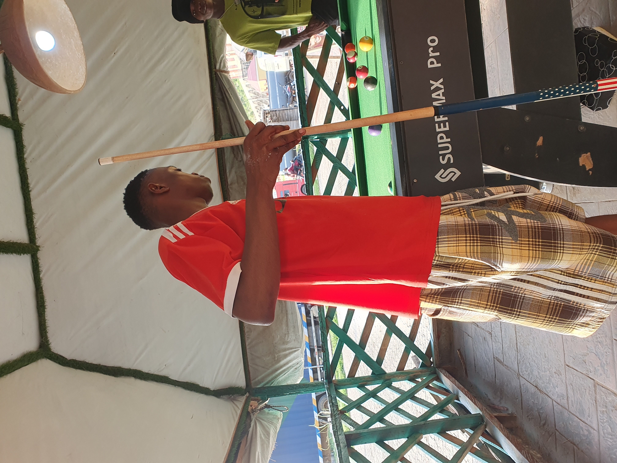 In Lokoja, Nigeria, a young Black male is captured in a medium shot, standing beside a green pool table and holding a billiard cue. He is dressed in a red t-shirt with white stripes on the sleeves and plaid shorts. The pool table, labeled "SUPER MAX Pro," has several balls on its surface. To the right of the frame, another Black male, wearing a green t-shirt and a black hat, is partially visible. The setting appears to be an outdoor or semi-outdoor space, with a white tent-like covering overhead and a wooden lattice structure in the background. The ground is paved with interlocking tiles. A truck is visible in the far background, suggesting the location is near a road or transport hub. The lighting suggests it is daytime.