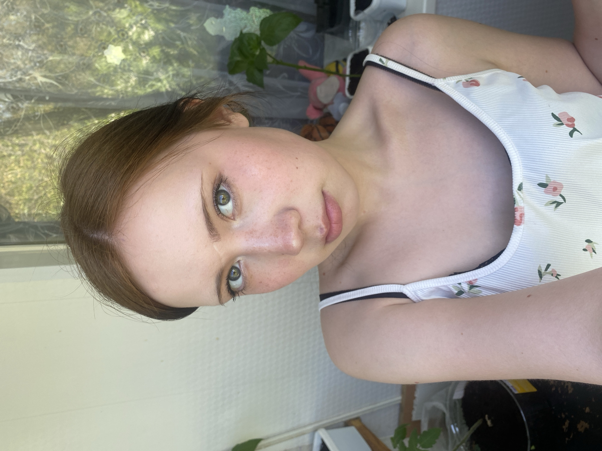 A light-skinned person with brown hair and green eyes is visible in this close-up portrait. The person is wearing a white tank top with a floral print and black straps. The background includes a window, a white wall, and indoor plants. The location is Unknown, Unknown.