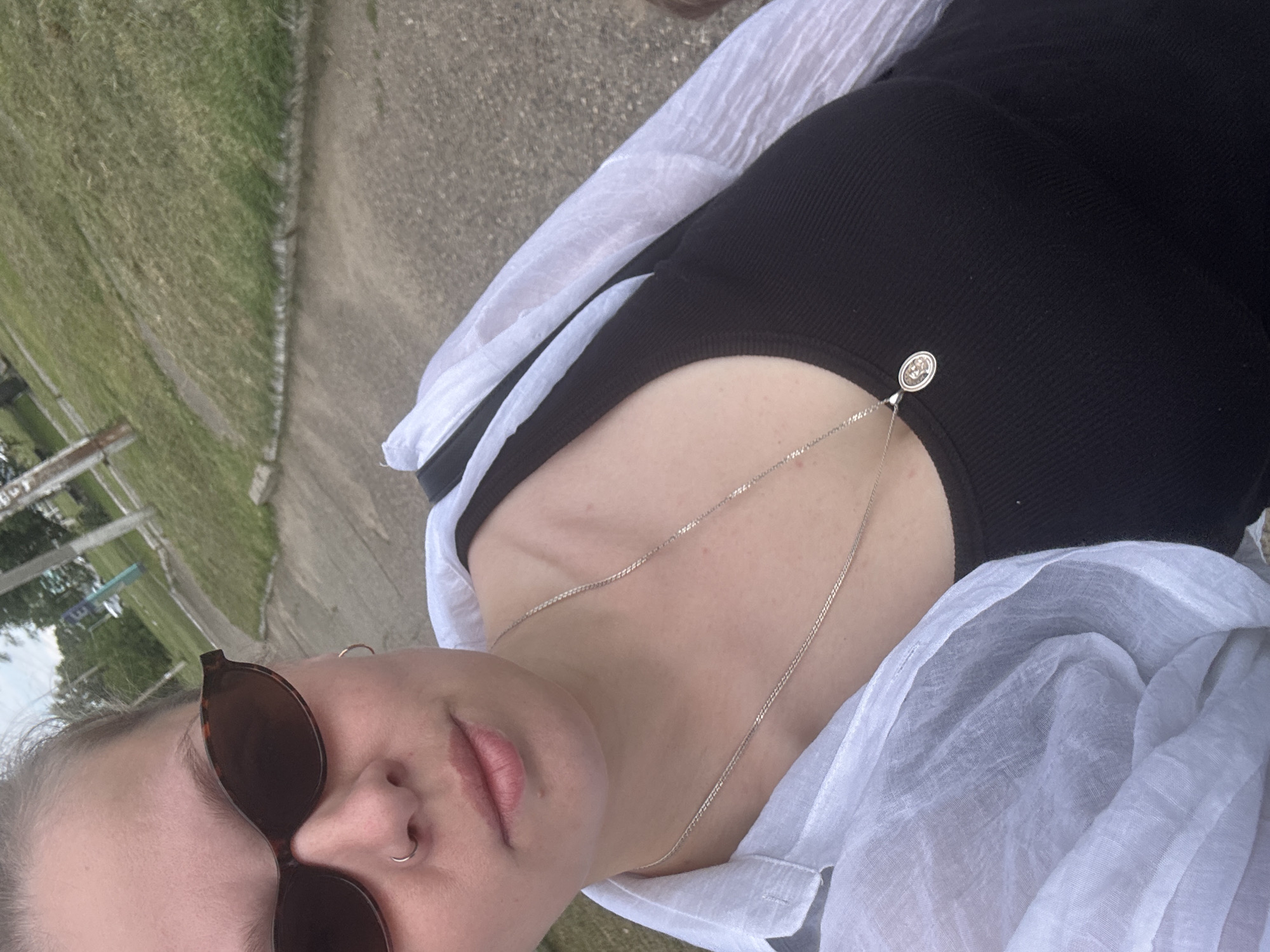 The media file is a close-up, low-angle, outdoor selfie taken by a person wearing sunglasses and a black top with a silver necklace. The person is also wearing a light-colored, sheer garment draped over their shoulders and chest. The background shows a grassy area and a paved path, suggesting an outdoor setting like a park or a sidewalk. The lighting appears to be natural daylight, possibly overcast given the lack of strong shadows. The person's expression is not fully visible due to the sunglasses, but the open mouth and slight tilt of the head could suggest a casual or relaxed mood. There is no visible text in the image. The provided location context is Rozhdestvenskaya, Russia.