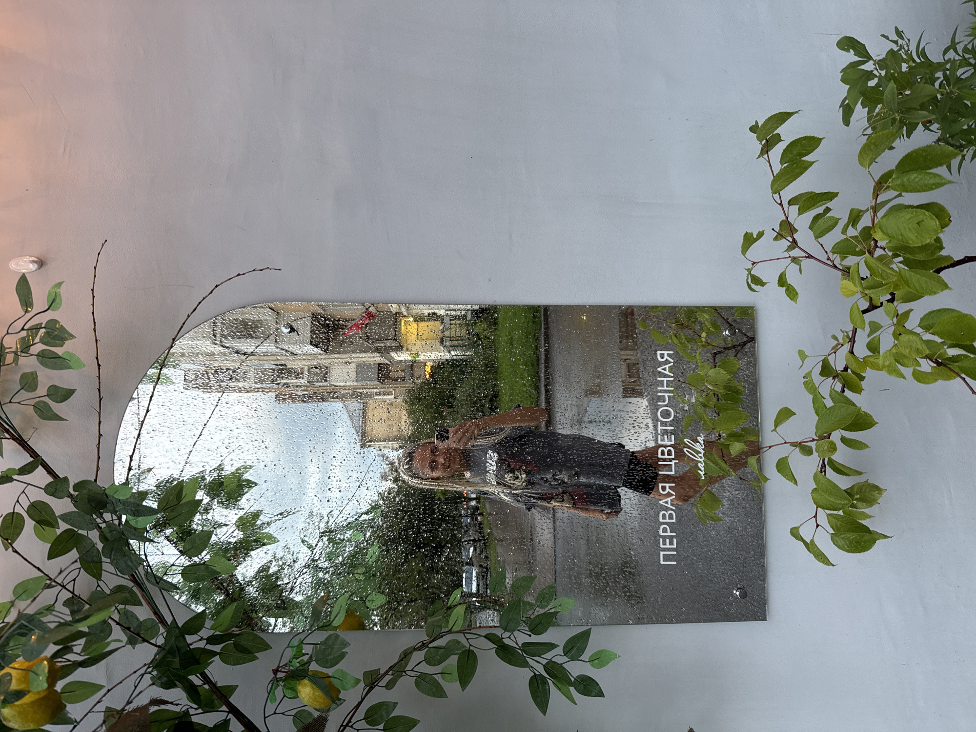 An arched mirror is affixed to a light-colored wall, with branches of a faux lemon tree arching over its top. The mirror reflects a street scene in what is stated to be Fedorino, Russia, under a cloudy, rainy sky. Water droplets are visible on the surface of the mirror, obscuring parts of the reflection.
In the reflection, a woman with blonde braided hair is taking a selfie with her phone. She is wearing a dark graphic t-shirt and black shorts. The background of the reflection shows apartment buildings, a street with parked cars, and green grass. Text is visible on the mirror itself, reading "ПЕРВАЯ ЦВЕТОЧНАЯ" (Pervaya Tsvetochnaya) and "lavka" in a script font below it.
In the foreground, to the right of the mirror, a live branch with green leaves extends upwards. To the left of the mirror, another branch of the faux lemon tree with green leaves and yellow lemons hangs down. The wall itself appears to be textured and painted a light color, possibly white or light gray. A small white circular object, possibly a light fixture, is visible on the wall above the mirror on the left. A small metal screw is visible on the bottom left of the mirror, securing it to the wall.
