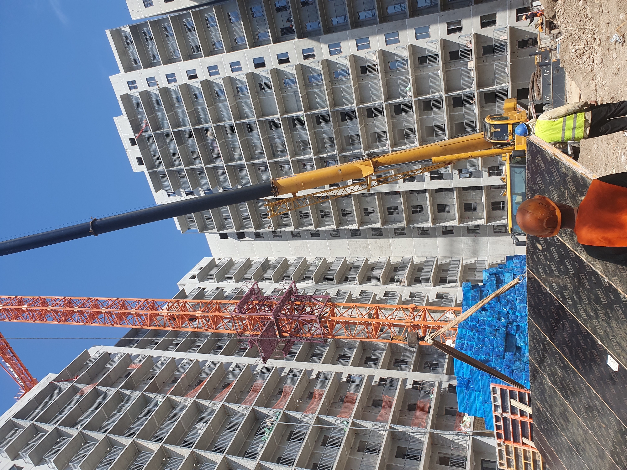 In Tashkent, Uzbekistan, a construction site features the skeletal framework of several high-rise residential buildings under construction. An orange tower crane stands prominently, its boom extended upwards, alongside a yellow mobile crane with its boom also raised. A load of blue-wrapped materials, likely construction supplies, is suspended by cables from the tower crane. Two construction workers, one wearing an orange hard hat and the other a blue hard hat and a yellow high-visibility vest, are in the foreground, facing away from the camera, with large wooden boards on the ground in front of them. Another worker in an orange outfit is visible further in the background. The buildings themselves are concrete structures with multiple balconies and visible window openings, indicating ongoing development. The sky is clear and blue.