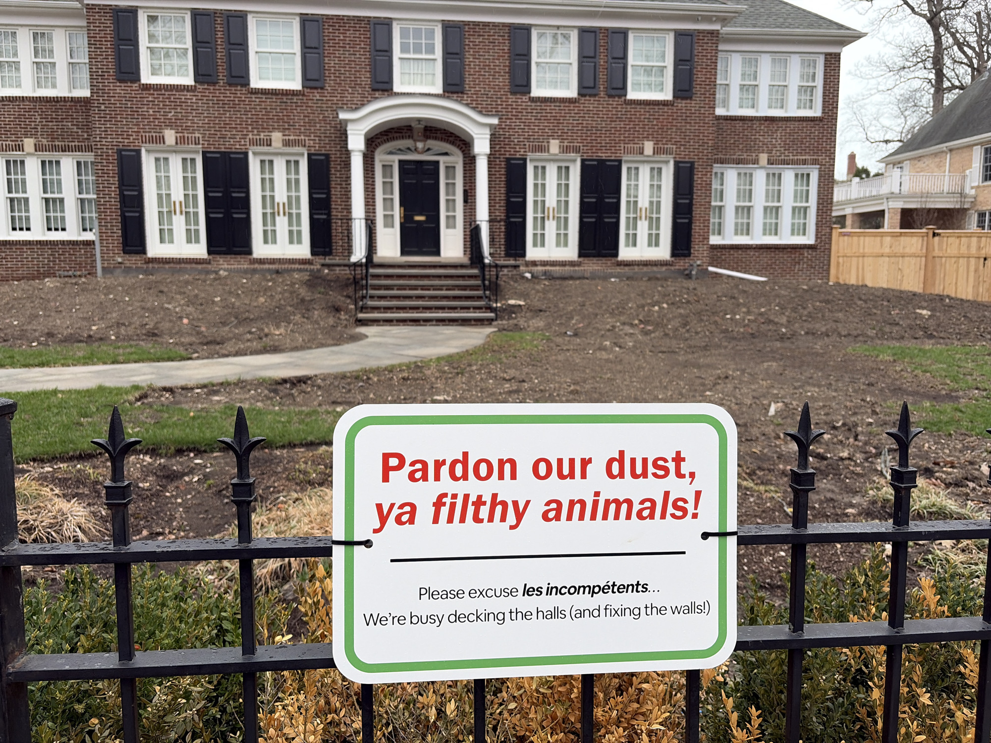 Here is a description of the image.
The image is a medium, eye-level shot of a sign attached to a wrought iron fence in front of a house. The house is a two-story brick building with black shutters and a white doorway and window frames. There is a freshly turned dirt plot in front of the house.
The sign reads "Pardon our dust, ya filthy animals! Please excuse les incompétents... We're busy decking the halls (and fixing the walls!)" The sign is white with a green border and black lettering. It's attached to the black wrought iron fence, which runs along a grassy sidewalk. Behind the fence is some shrubbery, some of which appears dead.
The picture was taken in Winnetka, United States of America.