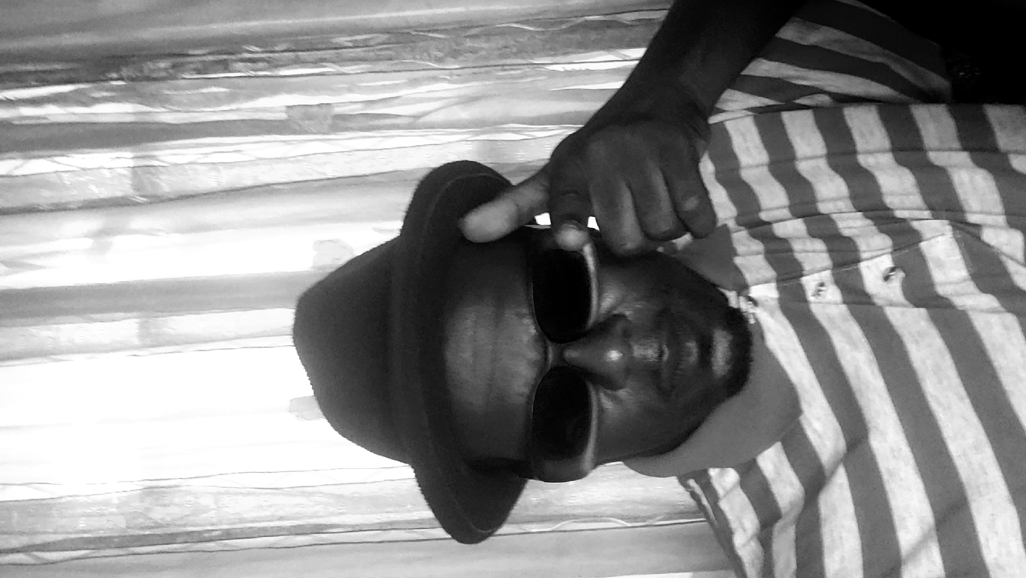A black and white close-up of a person wearing a hat and sunglasses, lying down on a striped shirt. A hand touches the hat. The background shows textured, possibly wrinkled, fabric. The location, Narayi, Nigeria, is not discernible from the image.