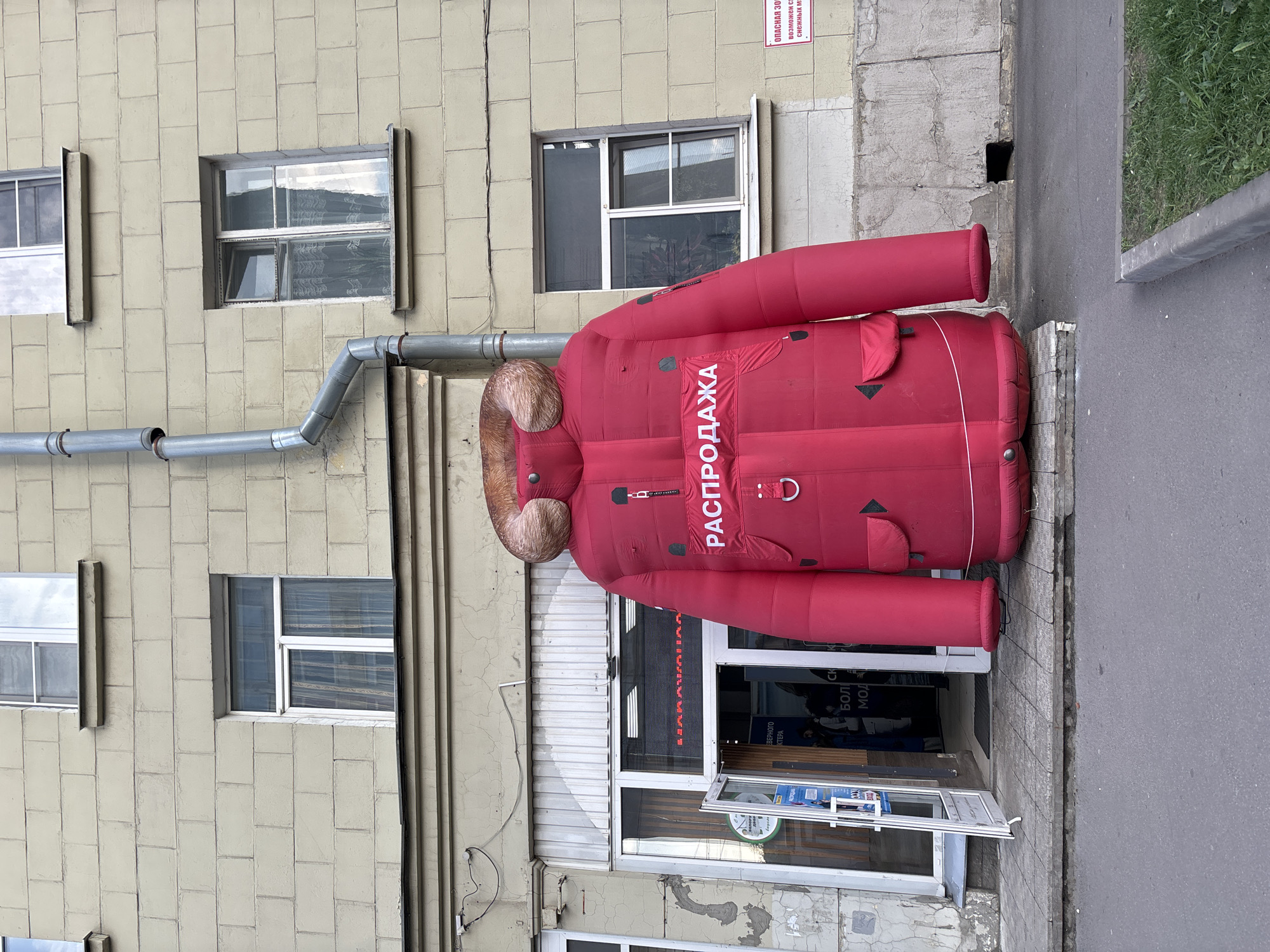 The image shows a red inflatable jacket advertising a sale ("РАСПРОДАЖА" in Russian) attached to the exterior of a building in an urban area. The building has a series of rectangular windows, a grey downspout, and a store entrance with an open door. The street is visible to the right of the building, with a section of curb and a patch of grass. The city is unknown, as no location indicators are present.