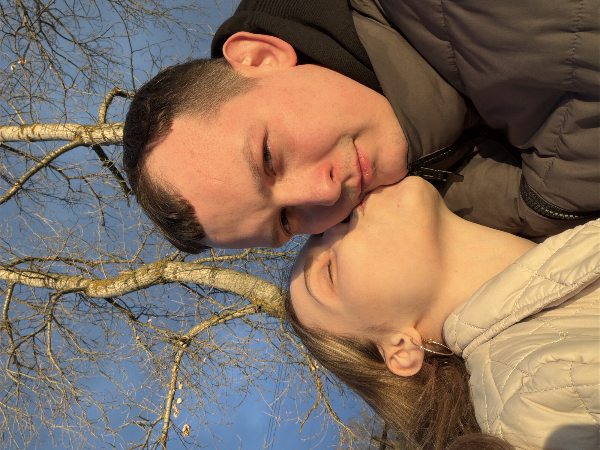 The image displays a close-up, intimate moment between two young individuals outdoors. The two subjects are positioned with their faces close together, oriented diagonally across the frame. One person, with short dark hair, wearing a black hoodie and a dark grey puffer jacket, has their eyes mostly closed with a subtle, contented smile. Their chin is gently met by the closed lips of the second person, who has long light brown hair, is wearing a beige puffer jacket, and a hoop earring in their visible ear. Both individuals have their eyes closed, conveying a sense of affection, tenderness, and happiness.
The background features a clear, bright blue sky and the intricate network of bare tree branches. The main visible tree trunk shows light-colored bark, indicative of a deciduous tree, possibly a birch. The absence of leaves on the branches suggests the photo was taken during late autumn, winter, or early spring. The lighting appears to be the golden hour, casting a warm glow on the subjects' skin and the tree, indicating either early morning or late afternoon. The puffer jackets worn by both individuals suggest cool or cold weather conditions, consistent with the likely climate in Chaikovsky, Russia, the provided location context. There is no visible text in the image.