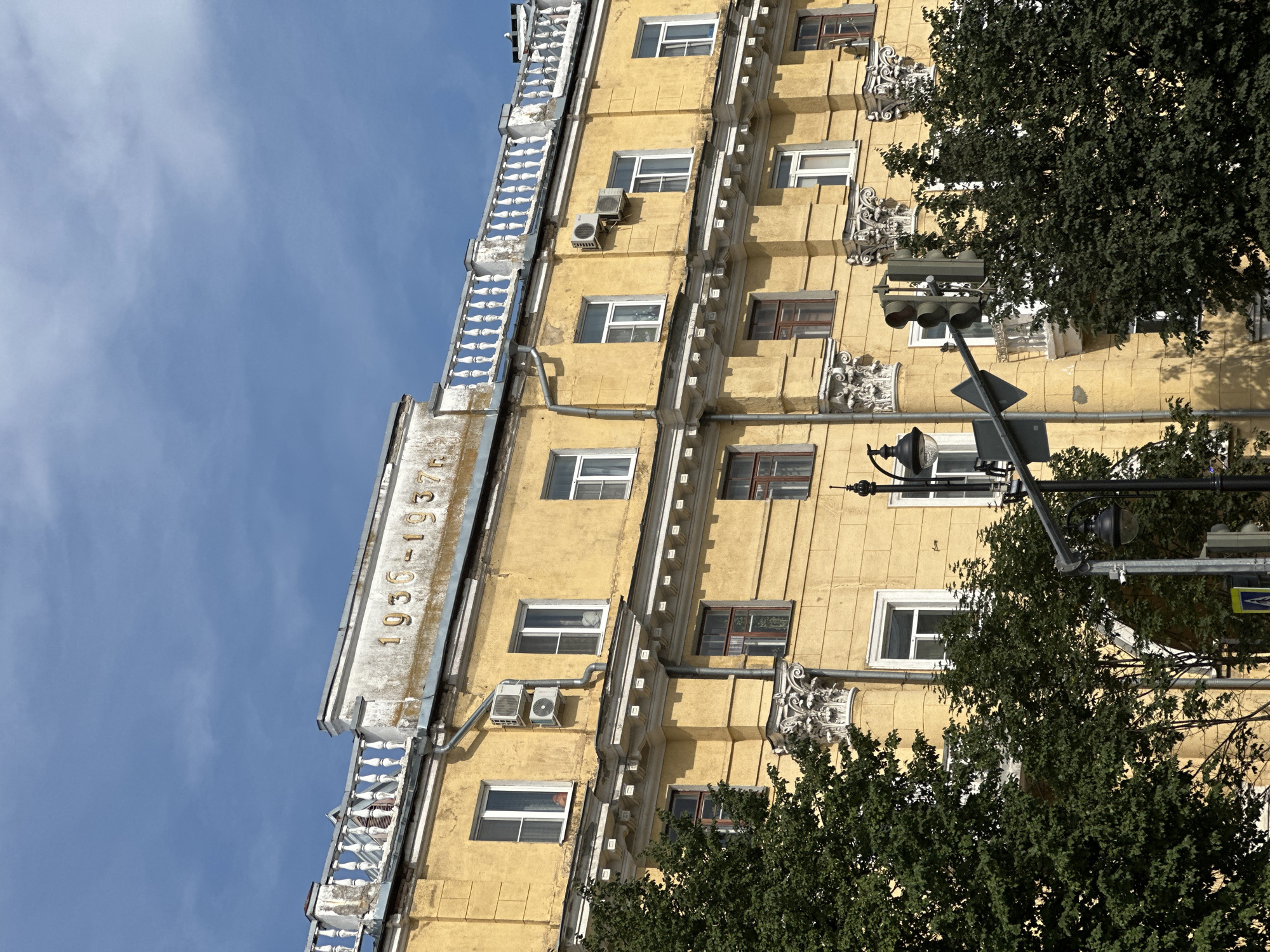 The image shows a yellow apartment building with the inscription "1936-1937" on its facade. The building has multiple windows, some with air conditioning units attached. There is a decorative railing along the top of the building. Trees are visible in front of the building, and a traffic light and street lamp are also present. The sky is blue with some clouds. This building is located on Voskresenskaya Street in Arkhangelsk, Russia.