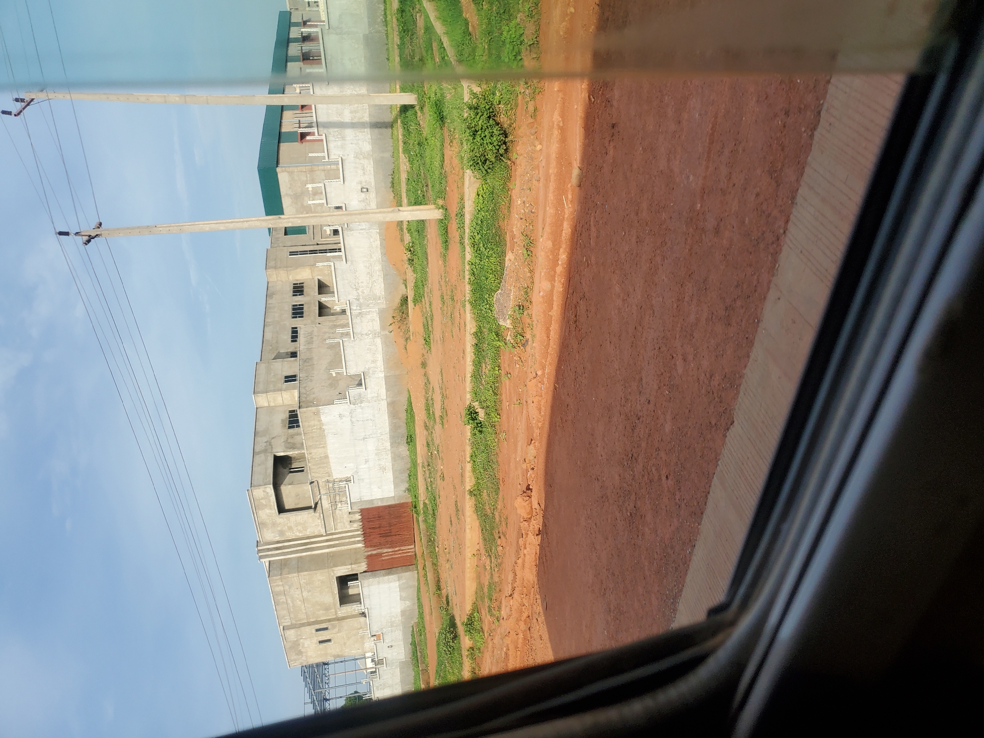 This is a medium-distance, eye-level shot of an ongoing construction project on a sunny day in Kakau, Nigeria. The scene is viewed from inside a vehicle, as evidenced by the dark window frame in the foreground. A partially completed building complex, constructed with concrete, is visible in the background. The building features a light-colored facade with a dark green metal roof on a section of it. A tall, rusty metal gate is situated in front of one of the building's entrances. Several utility poles with electrical wires are also present. The immediate surroundings of the building are cleared land with some patches of green grass. The foreground is dominated by a reddish-brown dirt road. The sky is blue with scattered white clouds.