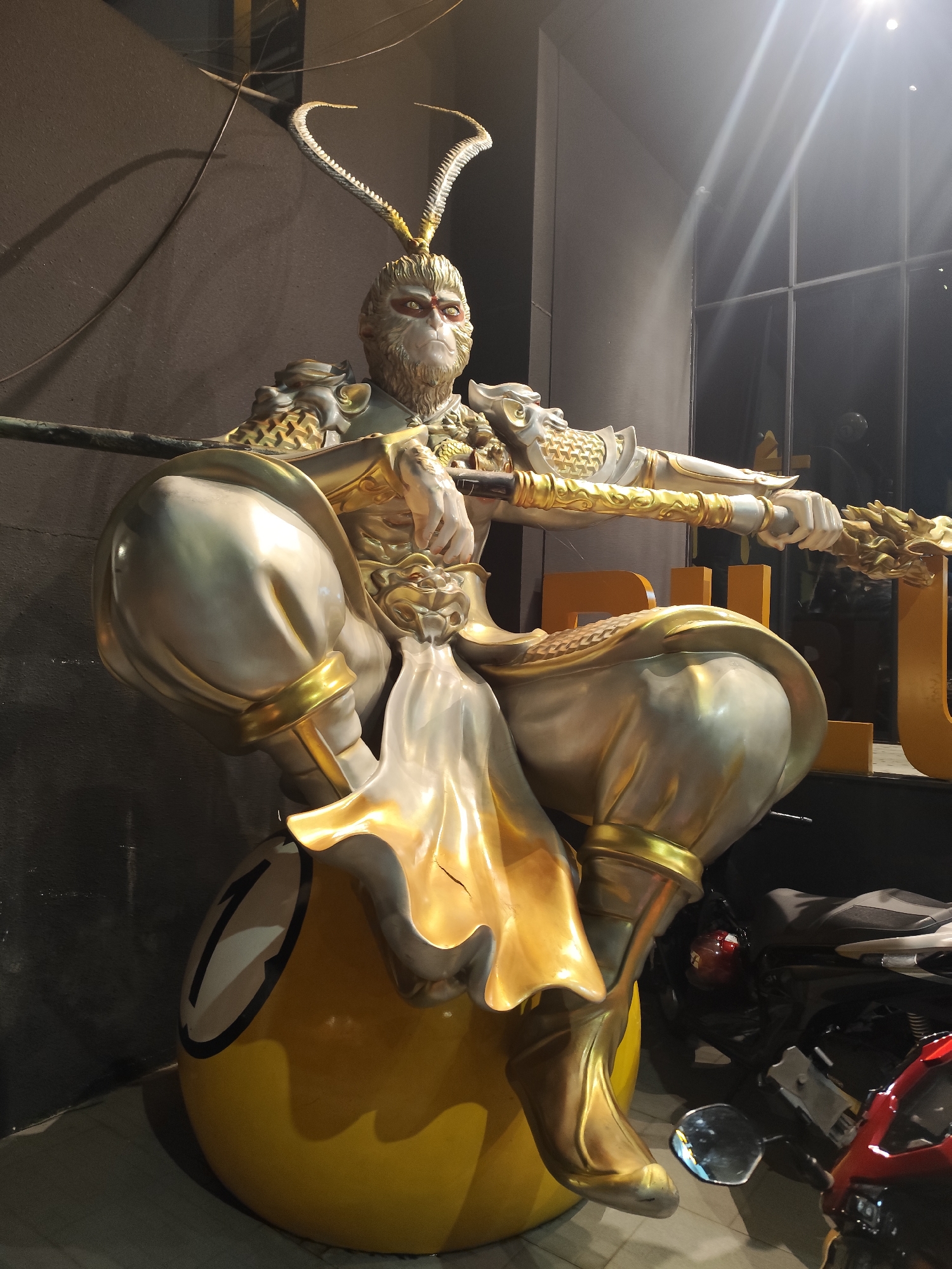 A large, ornate statue, resembling the Monkey King (Sun Wukong), is the central focus, depicted in a powerful squatting pose. The statue is predominantly gold and silver, adorned with intricate armor and a flowing golden sash. Its face is monkey-like, with distinct red markings around the eyes, conveying a serious and determined expression. Two prominent, curved, antler-like appendages rise from its head. The figure holds a long, golden staff horizontally across its body with both hands.
The statue is seated atop a large, glossy yellow sphere, which is clearly identifiable as an 8-ball from billiards, marked with a black circle and the number "8" in white.
The setting appears to be indoors, with a dark, textured wall forming the background on the left. To the right, a large window or glass panel reflects bright artificial light sources. Partially visible behind the statue's right side are golden block letters, with "U" or "JU" being discernible. On the floor to the right of the statue, parts of two motorcycles or scooters, one dark and one red, are visible. Overhead, faint wires or cables are present. The artificial lighting suggests an indoor setting, likely an establishment such as a themed shop or entertainment venue in Da Lat, Vietnam.