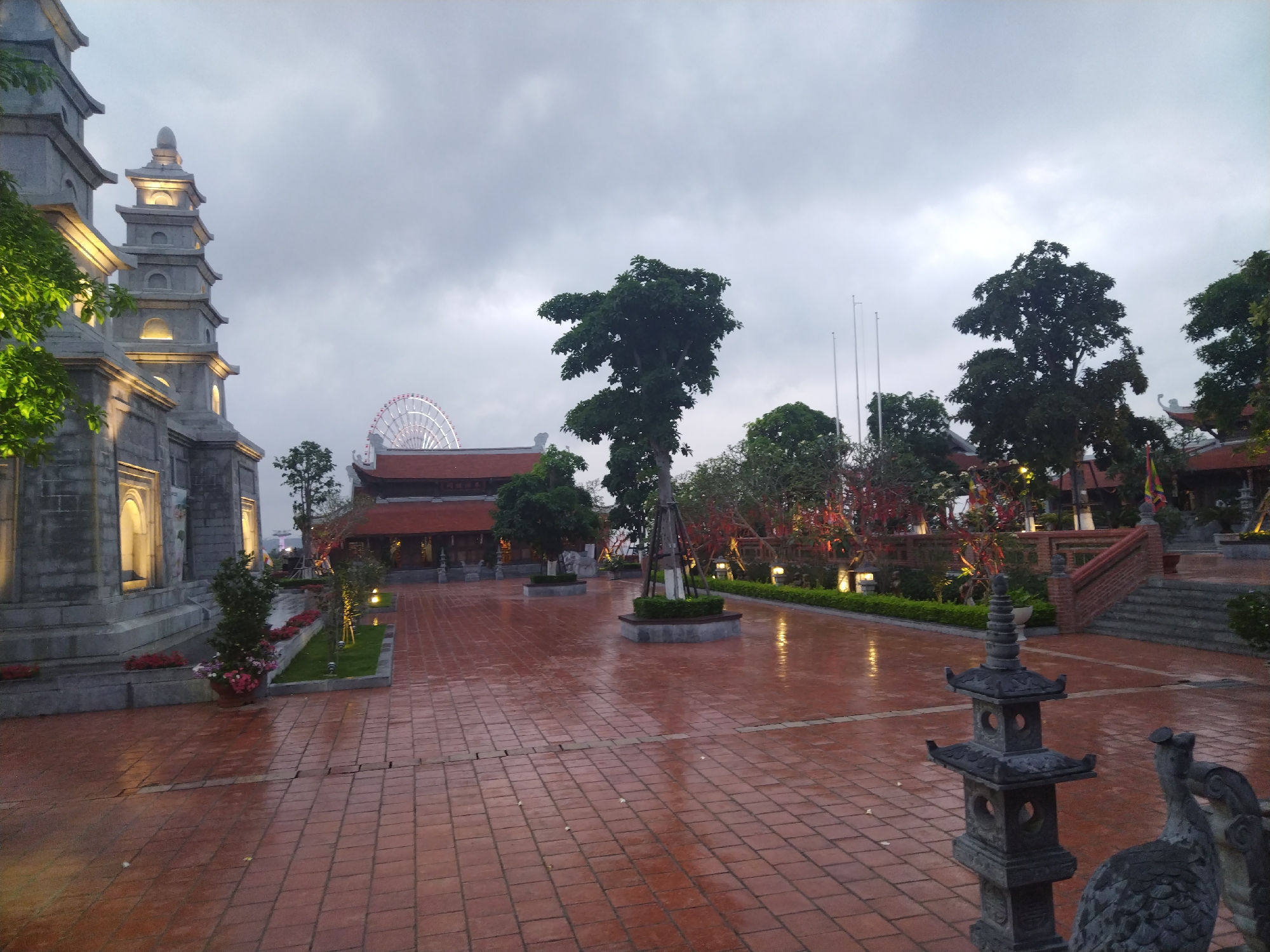 The image shows a wet, brick-paved plaza in Hong Gai Ward, Vietnam. In the background, there is a Ferris wheel. To the left, a multi-tiered stone structure is illuminated with golden lights. In the center of the plaza, there is a manicured tree. Other greenery, including shrubs and trees, are distributed around the plaza. The sky is overcast, and the light is muted. In the foreground, there is a stone sculpture of a pagoda-like structure and a bird.