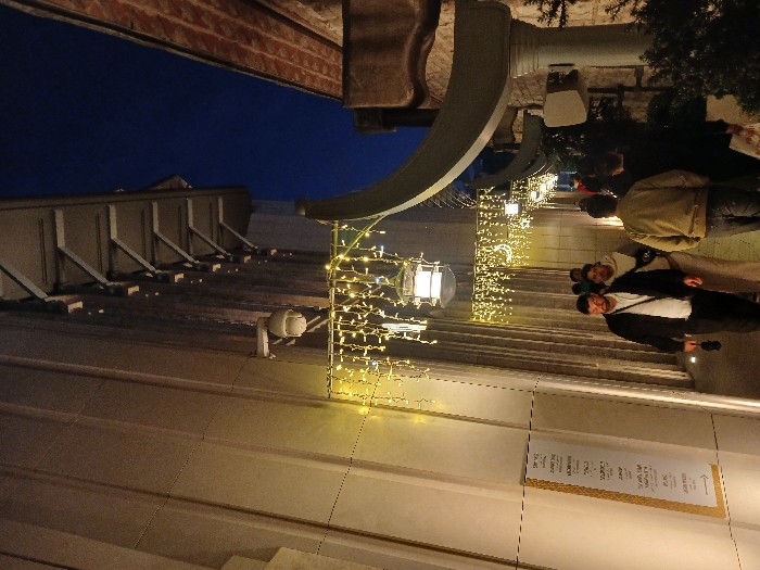 The image shows a narrow alleyway between two buildings in the Beyoğlu district of İstanbul, Türkiye. It is nighttime. String lights illuminate the alleyway. The building on the left is a modern, white building with a smooth, flat surface. The building on the right is older, with a rough, stone facade. The buildings are lined with strings of lights, and there are several people walking in the alleyway. The people are dressed casually, and some of them are carrying bags. The alleyway is empty except for the people. There is a sign on the wall of the building on the left that reads "Deniz". There is also a security camera mounted on the wall of the building on the left. The image is taken from the ground level, looking up towards the top of the alleyway.