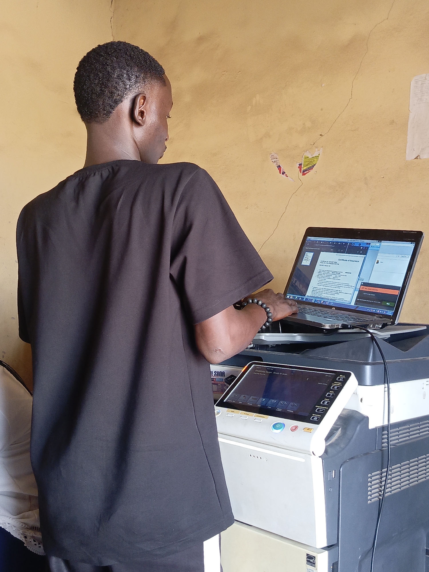 An individual, viewed from behind, stands operating a laptop placed atop a large multifunction printer or copier machine in what appears to be an indoor setting in Akwanga, Nigeria. The person, a young man with short, dark curly hair and dark skin, is wearing a loose-fitting black short-sleeved top and a beaded bracelet on his right wrist. He is leaning slightly forward, with his hands positioned over the laptop's keyboard, indicating active use.
The laptop screen displays what looks like a document or webpage with the prominent heading "Certificate of Insurance." Other text and user interface elements, including what might be a progress bar or status indicator, are visible on the screen. The laptop is a silver-colored model, possibly a Dell, and is connected to the printer via a cable.
Below the laptop, the large grey and white multifunction printer/copier features a control panel with a digital screen. On this screen, the text "Stabilizing Image. Ready to scan." is visible, alongside various touch-screen buttons and icons for different functions. A partial "SAMSUNG" logo can be seen on the side of the machine. Near the base of the printer, an "ENERGY STAR" logo is discernible.
The background consists of a plain, light brown or beige wall with visible cracks running vertically. Several torn pieces of paper or posters are haphazardly affixed to the wall, with some showing fragmented text and splashes of red and yellow colors. The overall impression is that of a functional workspace, possibly a small business or office, where printing and computer services are provided. The lighting suggests daytime.