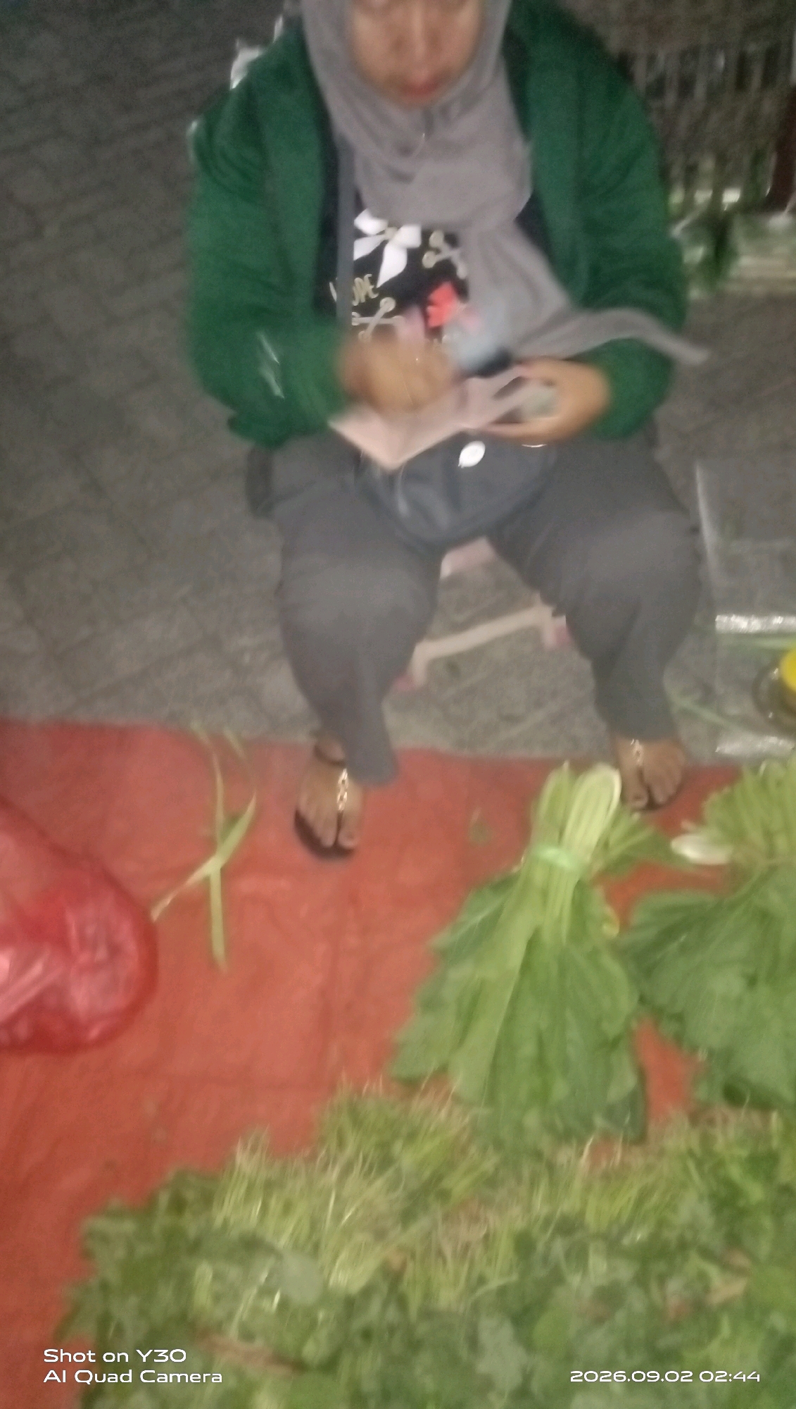 A person wearing a green jacket, grey hijab, and grey pants sits on the ground in Bulurejo, Indonesia. The person is holding a phone in their hands. In front of the person are bundles of green leafy vegetables and a red plastic bag. The scene appears to be a market setting.