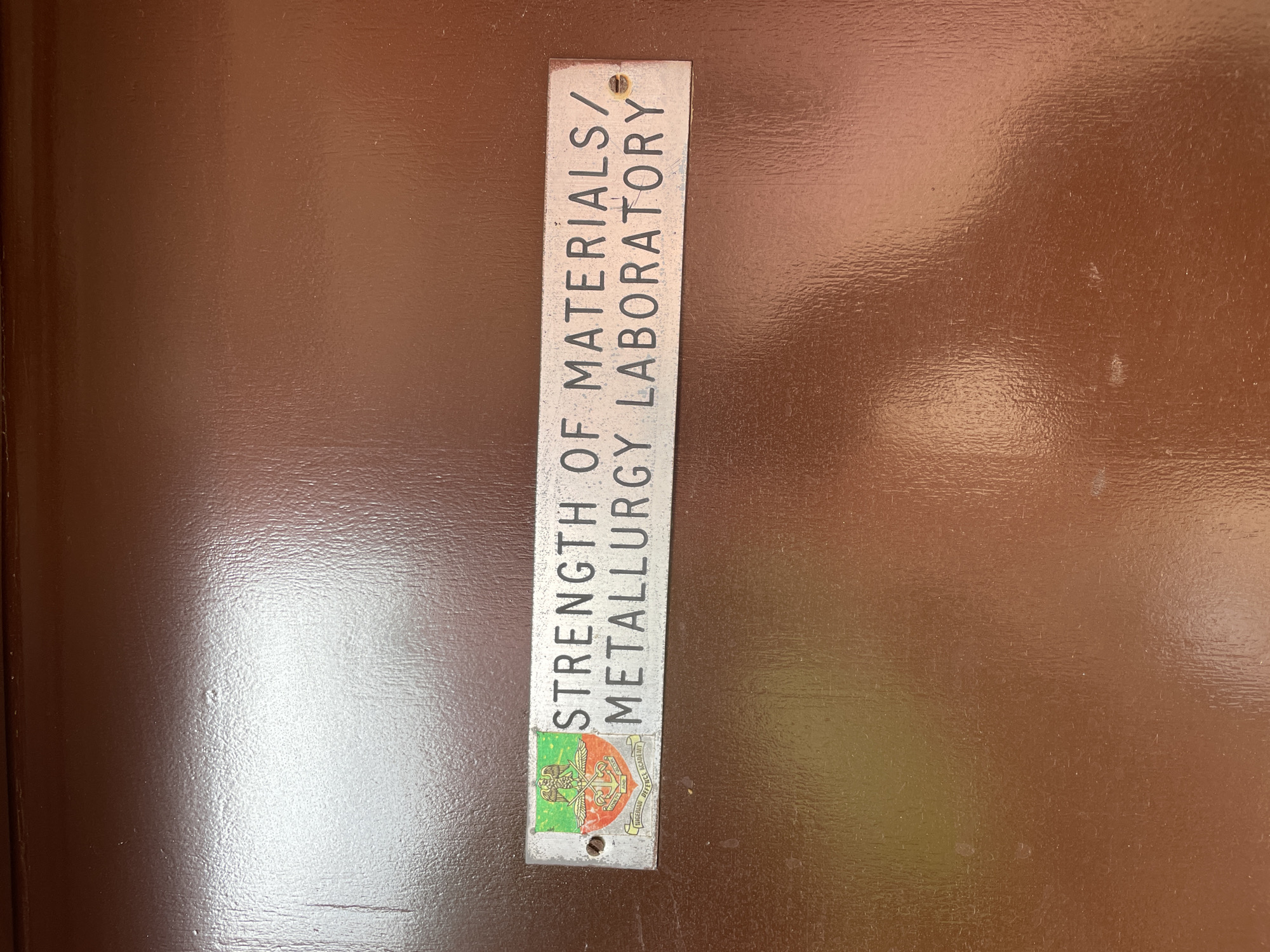 The image shows a metal sign affixed to a brown surface, likely a door or wall. The sign reads "STRENGTH OF MATERIALS/METALLURGY LABORATORY" in black lettering. Below the text is a colored emblem featuring a green field on one side and a crest that includes the words "NIGERIAN DEFENCE ACADEMY". Based on the crest, the location of the laboratory is likely within the Nigerian Defence Academy in Afaka, Nigeria.