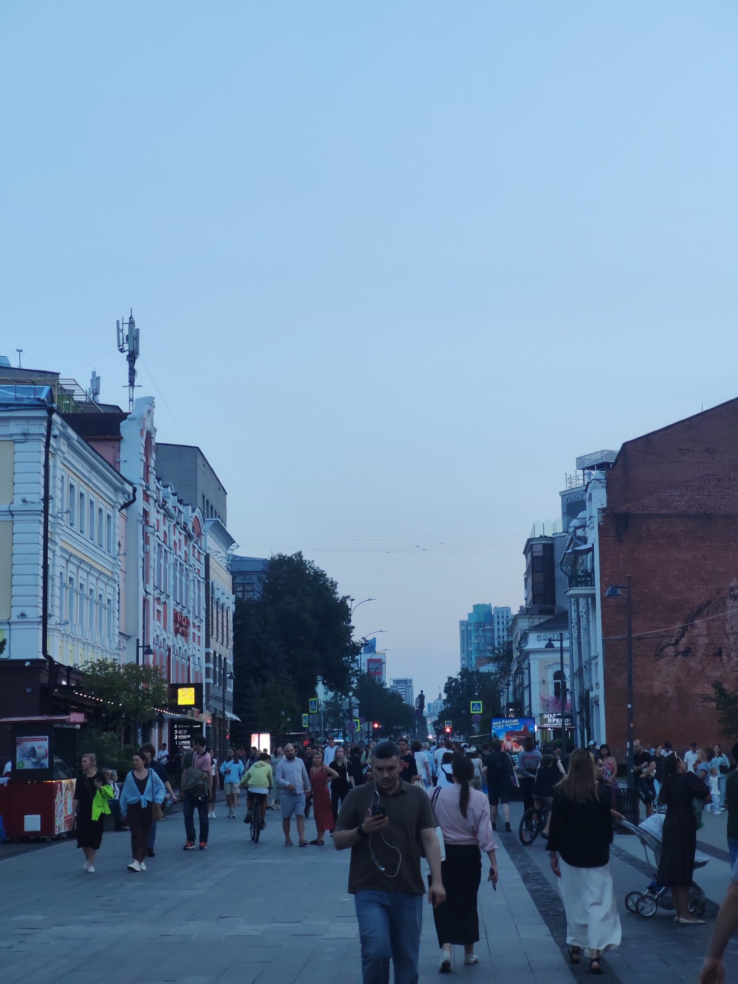 The image depicts a street scene in Nizhny Novgorod, Russia, likely during the evening. A wide pedestrian walkway is filled with numerous people walking, talking, and interacting. The architecture on either side of the street features multi-story buildings with distinct facades, some appearing historic and others more modern. Several businesses, including shops and possibly restaurants, are visible along the street. Traffic signals and streetlights are present, indicating it's an urban environment. The sky is a pale blue, suggesting twilight or early evening. A cellular tower is visible on top of one of the buildings. The overall impression is a lively, populated urban street.
