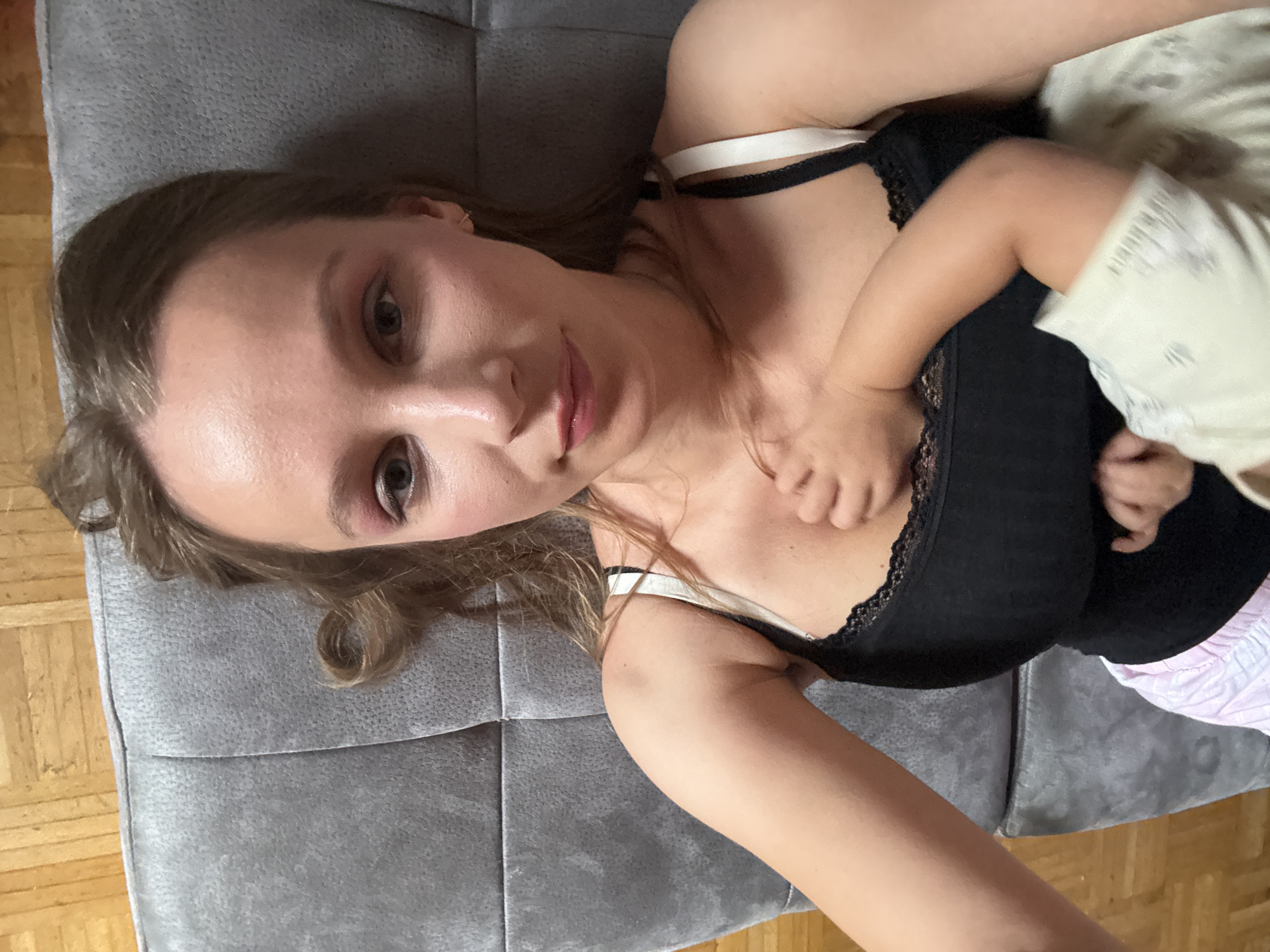 A woman, with blonde hair and visible makeup including pink eyeshadow, is lying down. She is wearing a black tank top with white straps and lace trim. A small child's hand is visible resting on her chest, with another part of the child's arm and clothing also in view. They appear to be indoors, on a grey, tufted couch, with a wooden floor visible in the background. This scene is likely within a residential setting. No specific location in Überherrn, Germany is indicated.