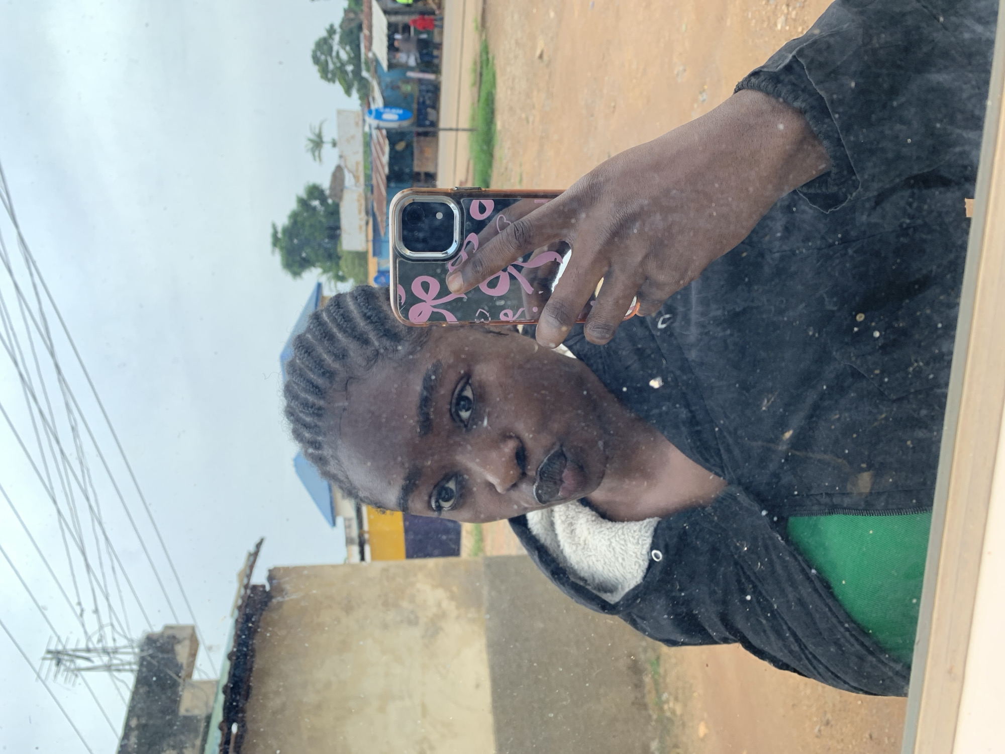 A young Black woman is taking a mirror selfie in what appears to be an outdoor setting in Jos, Nigeria. She has her hair braided in cornrows and is wearing a black jacket with a white furry lining over a green top. She is holding a smartphone with a pink bow pattern case to take the photo. The background shows a dusty, unpaved ground, a light-colored building to her left, and several structures with corrugated metal roofs and blue accents further in the distance. Power lines and poles are visible in the sky above. The mirror surface appears dirty, with specks and smudges that are reflected in the image.