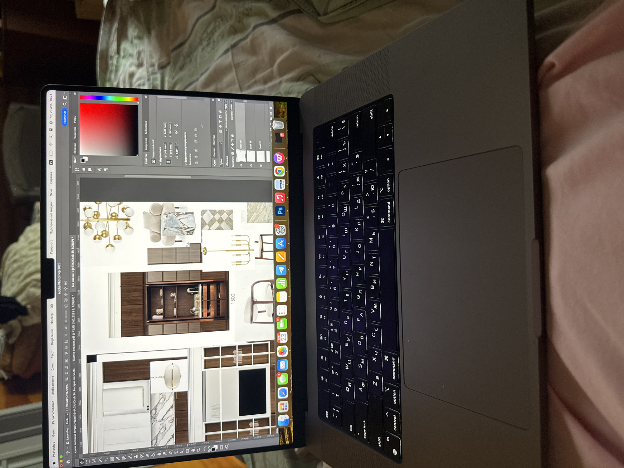 The media file captures a close-up, angled view of a MacBook Pro laptop placed on a light-colored, soft surface, likely a bed or sofa. The screen displays the Adobe Photoshop 2023 interface, open to multiple design projects.
The main activity shown is graphic design or interior design work. On the screen, two primary files are open: "кухня гостиная МУДБОРД.pdf" (kitchen living room MOODBOARD.pdf) and "Мастер спальня.pdf" (Master bedroom.pdf), indicating work on mood boards or design layouts for these rooms. One section of the screen shows a color picker with a red gradient selected, along with layers and properties panels. The design elements visible include modern gold-toned chandeliers, armchairs, a marble-patterned surface, various built-in cabinetry (some displaying decorative items), a white kitchen unit, and checkered floor tiles. Rulers with numerical markings are present around the design areas.
Notable details include the laptop's backlit keyboard, which features both Latin and Cyrillic characters, suggesting a Russian-speaking user. The menu bar at the top displays the time and date as "Чт, 23 апр. 16:24" (Thursday, April 23, 4:24 PM). The laptop's dock shows a variety of applications, including Adobe creative suite programs (Photoshop, Illustrator, InDesign), Microsoft Office applications, web browsers, and messaging apps (Telegram, WhatsApp, Viber). The background is indistinct but appears to be indoor, possibly a bedroom. The overall context, including the Cyrillic text on the screen and keyboard, aligns with the provided location of Tomilino, Russia.