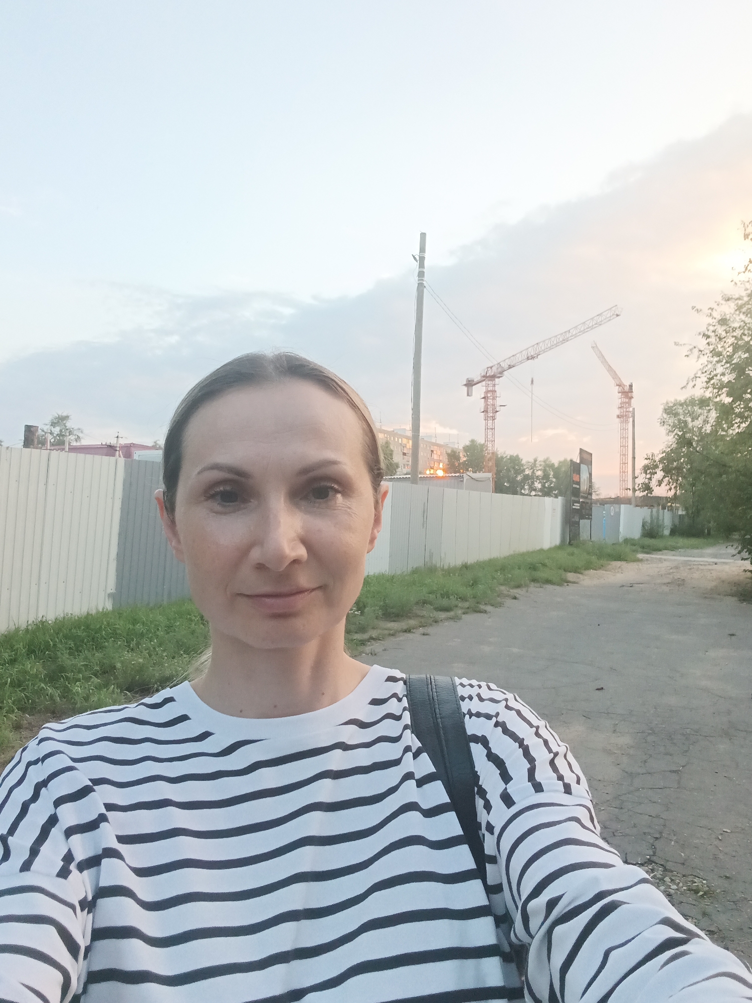 An outdoor selfie is taken by a woman with blonde hair, wearing a black and white striped long-sleeved shirt. She is standing on a paved path with a grassy area to her left and a corrugated metal fence behind her. In the background, two large construction cranes are visible against a hazy sky, suggesting a construction site. The setting appears to be an urban area in Komsomolsk-on-Amur, Russia, possibly a developing district. The lighting suggests it is late afternoon or early evening.