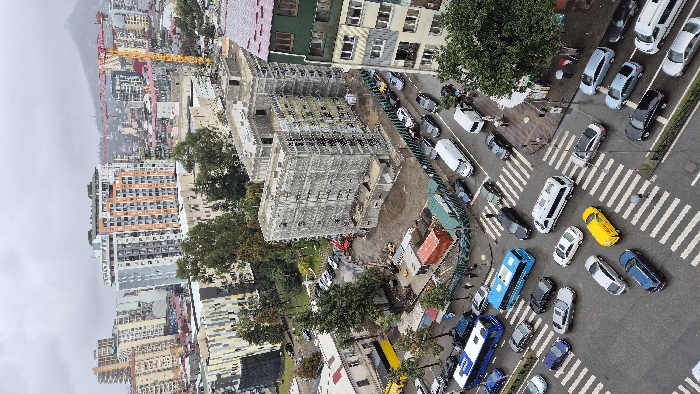This high-angle urban landscape image depicts an intersection and surrounding developments in Batumi, Georgia, under an overcast sky. The road surfaces are wet, displaying white pedestrian crosswalk markings. Traffic at the intersection includes two blue city buses, a yellow DHL van, and numerous private vehicles (sedans, SUVs, vans) of various colors, many appearing stationary. Pedestrians are observed on sidewalks and traversing crosswalks; some are equipped with umbrellas.
A multi-story concrete building under construction, partially scaffolded and with exposed concrete, occupies a prominent position near the intersection. Its perimeter is secured by a green corrugated metal fence, enclosing construction materials, red and green shipping containers, and a small red excavator. An unpaved lot adjacent to this site holds additional parked cars.
The broader urban environment features a dense array of multi-story buildings, including modern residential structures with distinct facades (e.g., white, grey, and orange sections; yellow and white stripes). A large yellow tower crane is visible against the horizon, signifying ongoing construction. Scattered trees are present throughout, and distant hills are discernible beneath the cloudy sky.