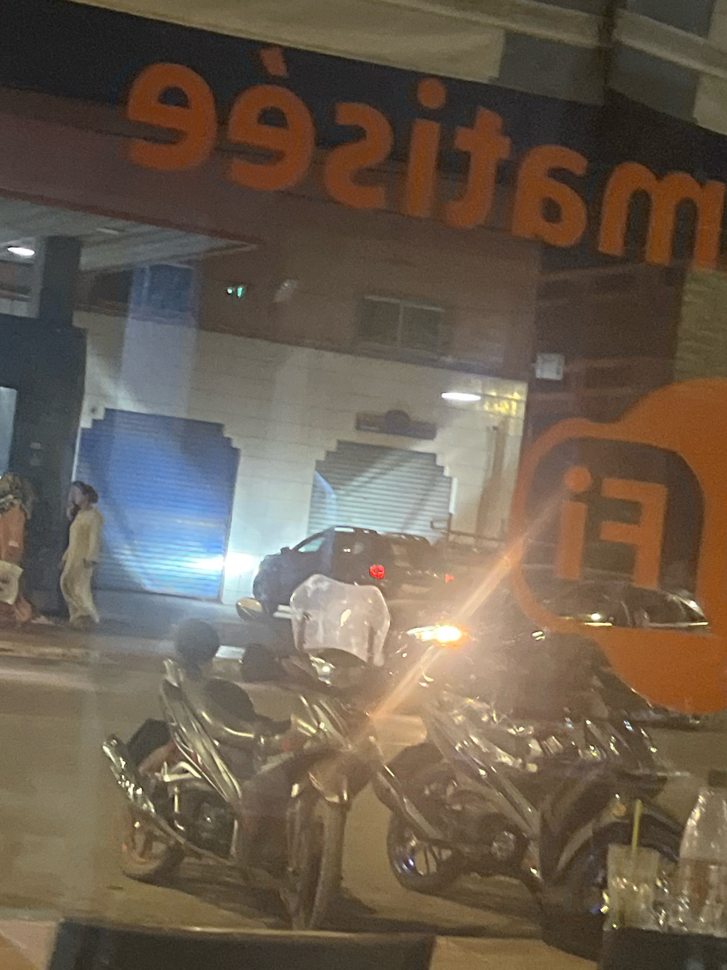The image depicts a street scene at night, likely in Azilal, Morocco, with a prominent orange sign featuring the text "992sit6m" in large, stylized lettering above a darker background. In the foreground, two motorcycles are parked, their headlights illuminated, casting a glare. One motorcycle is closer to the viewer and appears darker, while the other is slightly further back and more visible.
Behind the motorcycles, a dark-colored car is parked. To the left of the car, a person wearing a light-colored, flowing garment, possibly a djellaba, is walking away from the camera. Further to the left, another individual is partially visible.
The background consists of buildings with closed rolling shutters, one of which is a bright blue, and the other a lighter gray. The overall impression is a commercial area at night, with reflections visible on the surfaces, indicating a shopfront or display window. The presence of the motorcycles and the car suggests an active street or parking area.