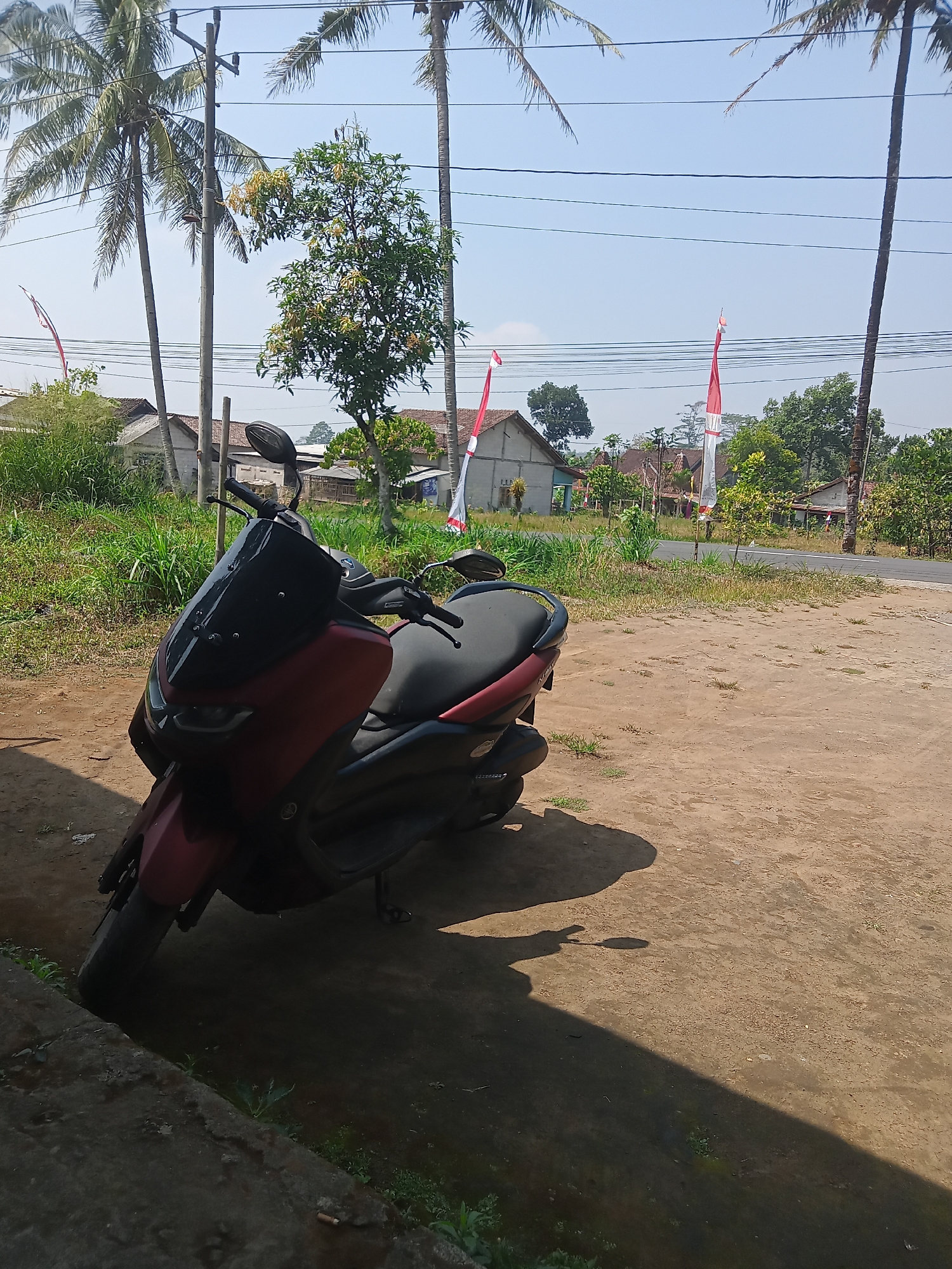 In Ngadirojo, Indonesia, a red and black scooter is parked on a dirt path next to a concrete curb. The scooter is the primary subject, positioned at a slight angle facing left. Behind the scooter, a road stretches into the distance, bordered by a grassy verge. In the background, several houses with red roofs are visible amongst lush greenery and tall palm trees. Indonesian flags, red and white, are mounted on poles and wave gently. The sky is clear and blue. The scene is illuminated by bright sunlight, casting shadows on the ground. There are no people visible in the image.