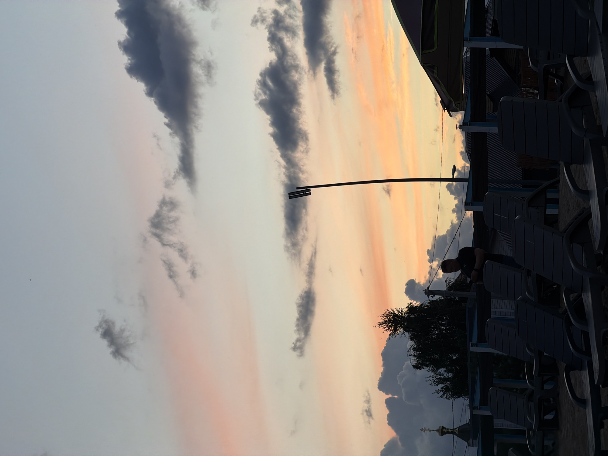 A vertical, outdoor, eye-level shot captures a dusky sky in Laishevo, Russia. Wispy grey and pink clouds streak across the horizon, illuminated by the warm glow of sunset. A silhouette of a person leans against a lamppost, their form cast against the vibrant sky.
In the foreground, rows of dark, empty lounge chairs are arranged outdoors, suggesting a public space like a cafe or beach area. The chairs are made of plastic and are in a clean, orderly fashion. Beyond the chairs, a dark silhouette of trees frames the scene, with a hint of a building with a golden dome peeking through the foliage in the lower left corner. The overall mood is peaceful and serene, with the quiet solitude of the chairs contrasting with the dramatic beauty of the sunset. The time of day is clearly evening, as indicated by the sunset colors in the sky and the silhouetted figures. No visible text is present in the image.