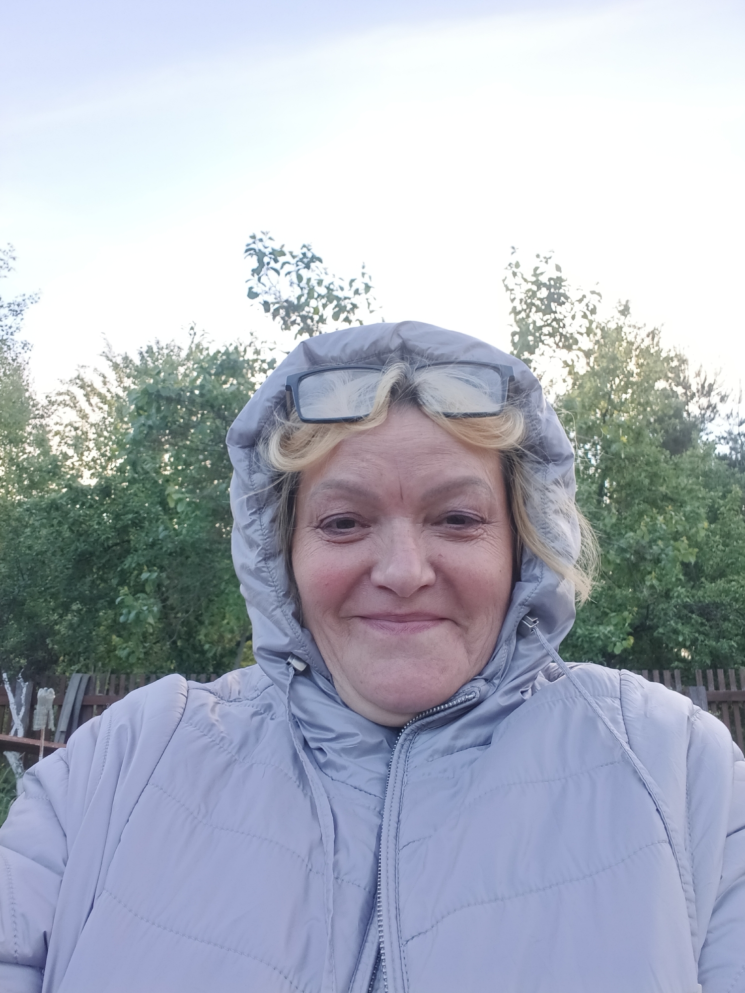 A selfie captures a middle-aged woman in Красный Маяк, Russia. She is wearing a light grey padded jacket with the hood up, with eyeglasses perched on her forehead above her blonde hair. The background includes green trees and part of a wooden fence, suggesting an outdoor setting. The sky is light and appears overcast.