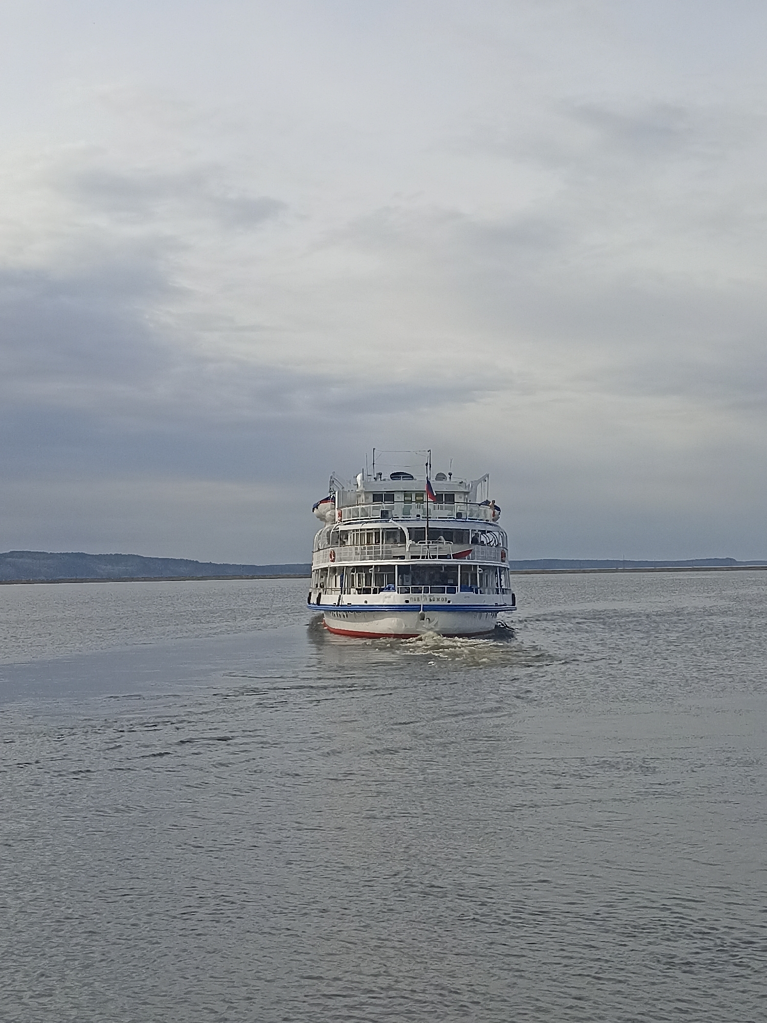 A cruise ship is moving away from the camera, leaving a wake in the water. The ship is white with red and blue accents and multiple decks. The sky is overcast with gray clouds. The location is Unknown, Unknown.