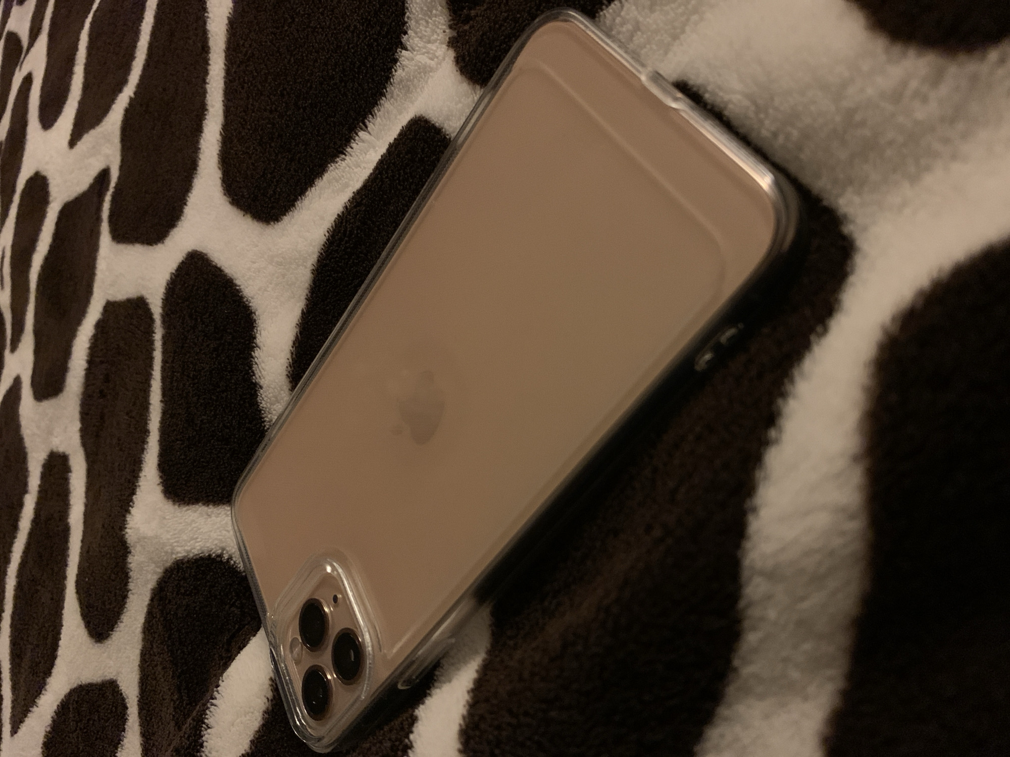 A rose gold iPhone 11 Pro in a clear case lies diagonally on a brown and white cow-print blanket. The phone is positioned with the rear camera array towards the top left corner of the image. The Apple logo is faintly visible on the back of the phone. The blanket has large, irregular dark brown patches on a white background. The lighting is dim, casting shadows. The image was taken indoors, likely in a domestic setting. There is no information available to determine the specific location within Чыжэвіцкі сельскі Савет, Belarus.