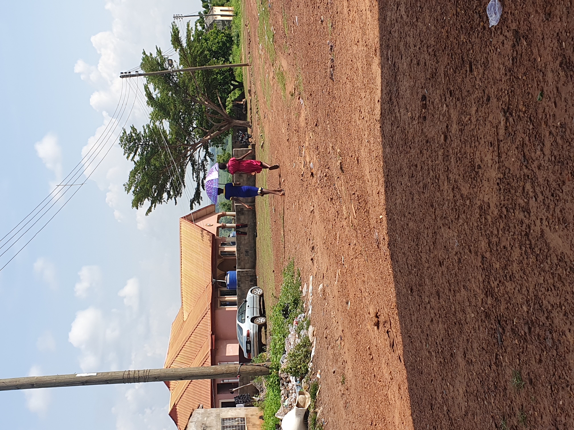 The image, rotated 90 degrees clockwise, captures an outdoor scene in what appears to be a semi-urban or rural area of Idah, Nigeria, under bright daylight.
The foreground is dominated by a wide, unpaved reddish-brown dirt path or ground, showing textures and scattered debris including plastic waste. In the midground, two individuals are walking along this path. One person, dressed in a red outfit, walks slightly ahead of another person wearing a blue outfit. The person in blue holds a purple and white umbrella open, suggesting protection from sun or rain.
To the left of the path, several buildings with distinctive reddish-brown corrugated metal roofs are visible. A white car is parked near one of these buildings. A large tree with dense green foliage stands behind the walking individuals and buildings, casting some shade.
Utility poles with multiple power lines cut across the upper left portion of the frame, set against a bright blue sky dotted with fluffy white clouds. The overall impression is a sunny day in an area with a mix of natural elements, residential structures, and some signs of litter. No discernible text is visible within the image.