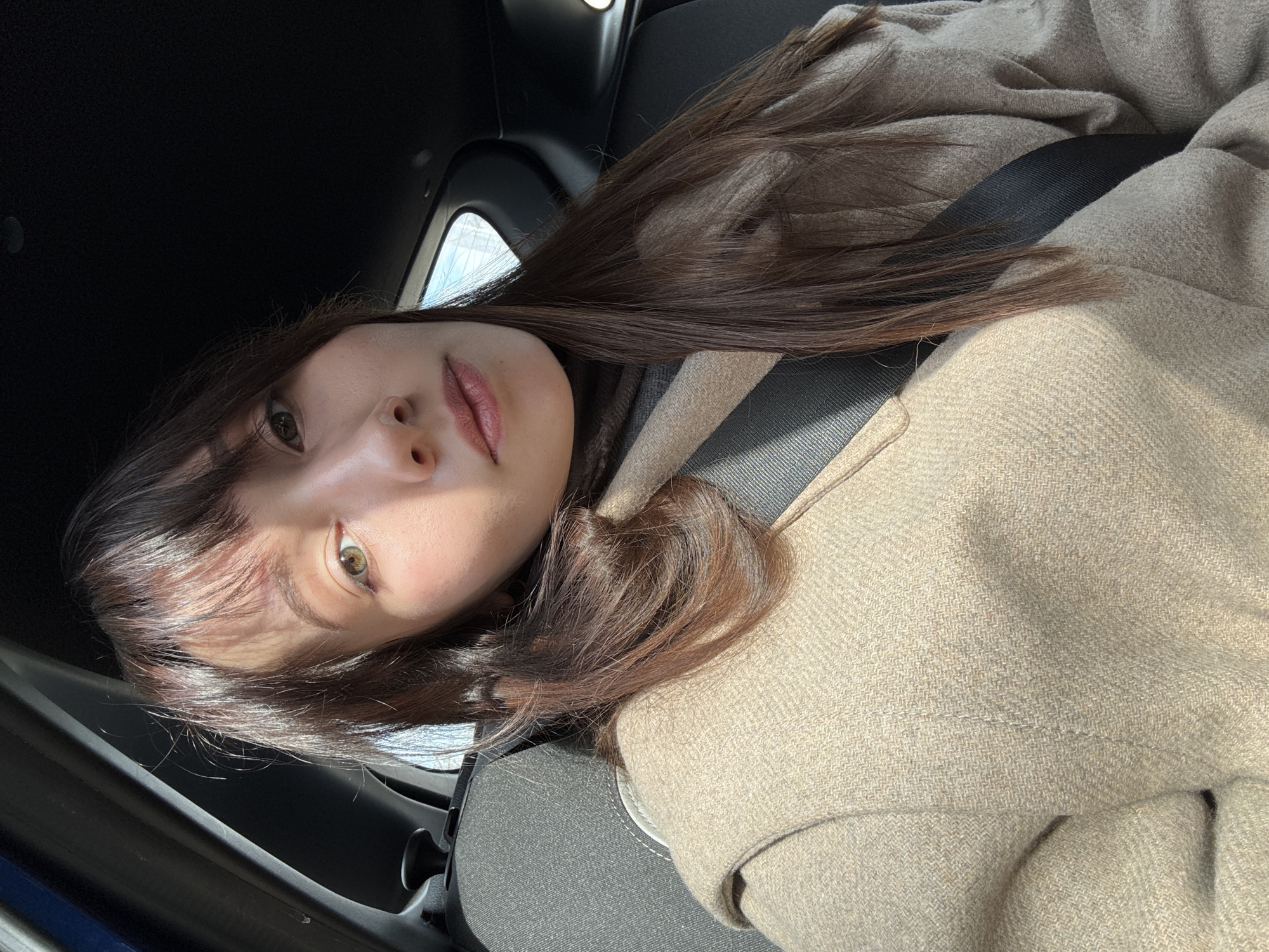 A woman with light skin, dark hair with bangs, and light eyes is sitting in the driver's seat of a car. She is wearing a beige coat and a seatbelt. The car's interior is black. The location cannot be determined to be in Kazan, Russia, based solely on the image provided.