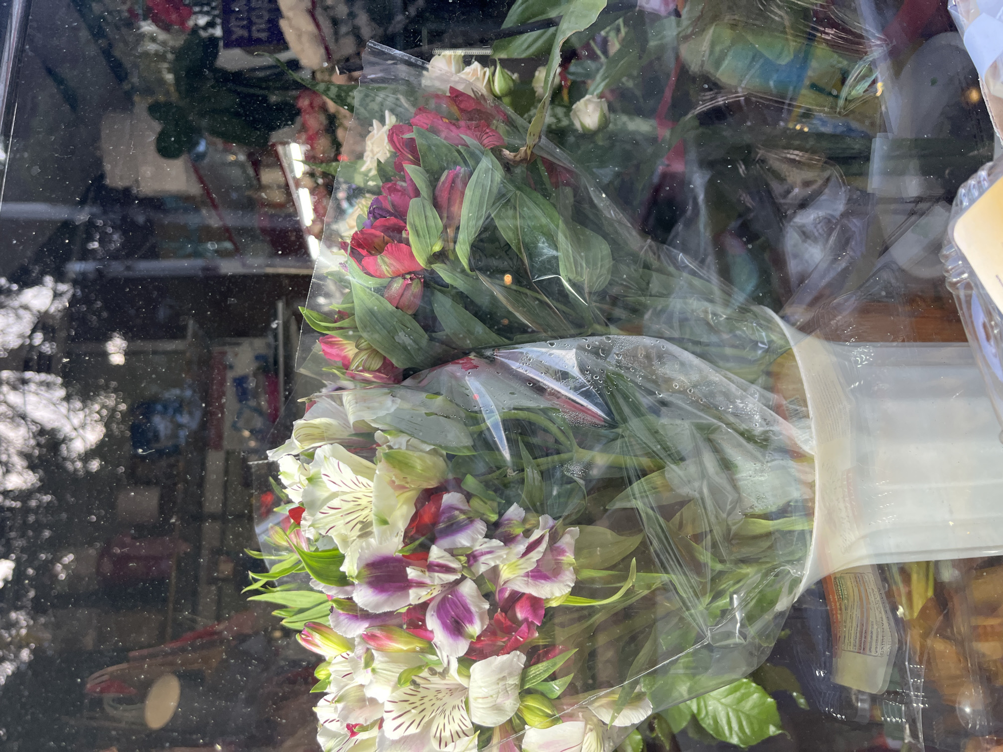 The image captures multiple bouquets of fresh flowers, primarily alstroemeria (Peruvian lilies), displayed behind a reflective glass surface, likely a shop window or a display case. The flowers are vibrant, showcasing hues of white, purple, red, and burgundy, with prominent green foliage and stems. Each bouquet is individually wrapped in clear plastic, which exhibits some condensation or water droplets.
The glass surface, slightly smudged with dust or dirt specks, strongly reflects both an outdoor scene and an indoor environment. On the left side, blurry reflections of what appear to be trees or bright foliage suggest a sunny, outdoor daytime setting. The right side and background show indistinct shapes and colors of an interior space, potentially shelves or other products within a store. Part of a yellow label on a packaged item is visible on the far right.
The scene depicts flowers arranged for sale, possibly in a florist shop or a grocery store in Sochi, Russia, according to the provided context. There are no people visible, and the flowers appear fresh and well-maintained. Any visible text on reflected signs or labels is too blurry to be deciphered.
