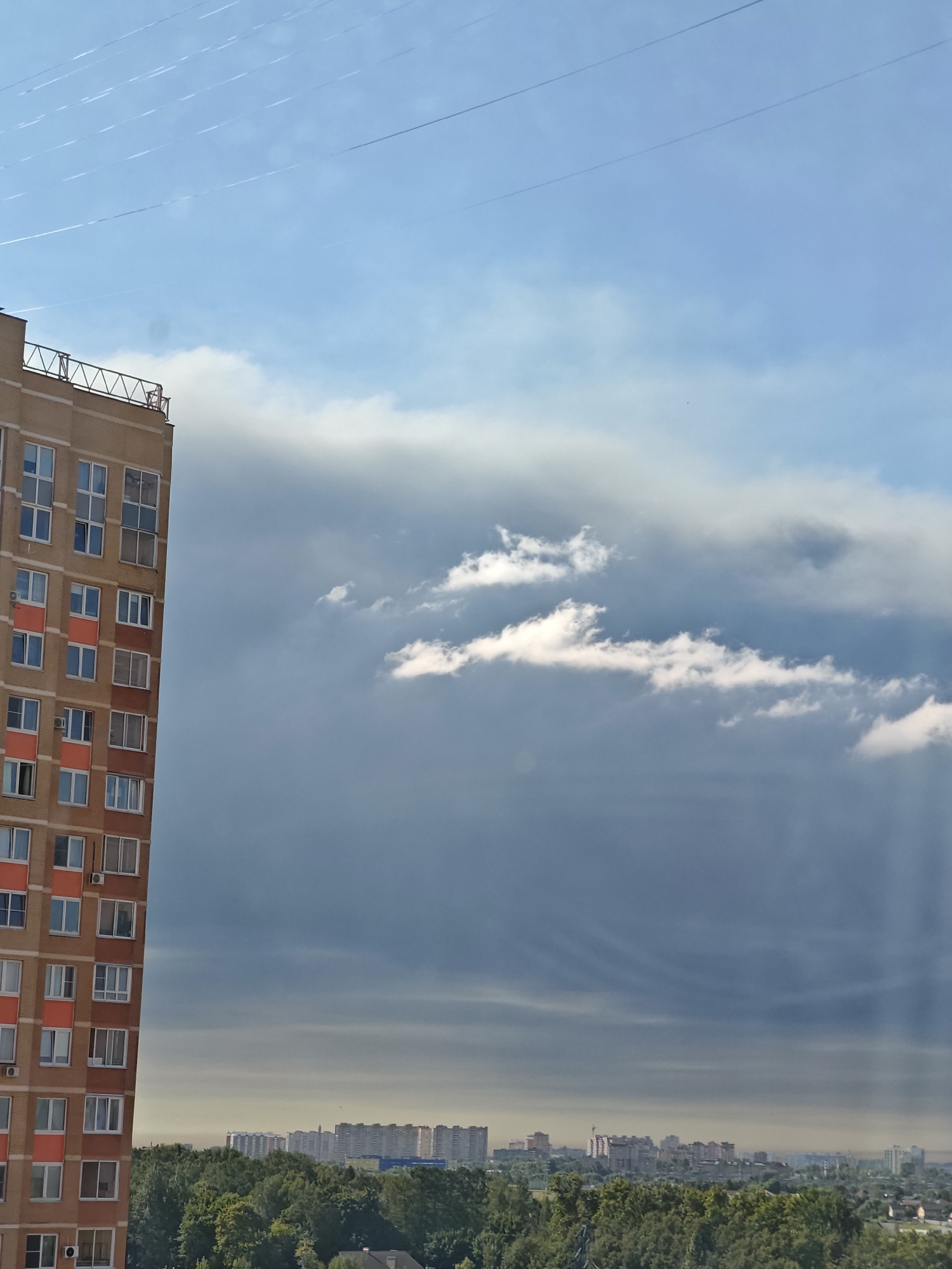 The image captures a cityscape under a dramatic sky. In the foreground on the left, a tall, multi-story apartment building with a brick facade and orange accents stands prominently. The building features numerous windows with white frames. Above the apartment building, several thin, white lines stretch across the blue sky, possibly contrails from aircraft.
Dominating the middle ground and extending into the background is a vast expanse of clouds, with darker, stormier clouds forming a dense mass, intersected by rays of light suggesting a break in the storm. Towards the horizon, a distant cityscape is visible, featuring a dense collection of buildings, including apartment blocks and other structures, interspersed with green trees. The overall atmosphere is one of an urban environment on a day with variable weather conditions. The image was taken in Shcherbinka, Russia.