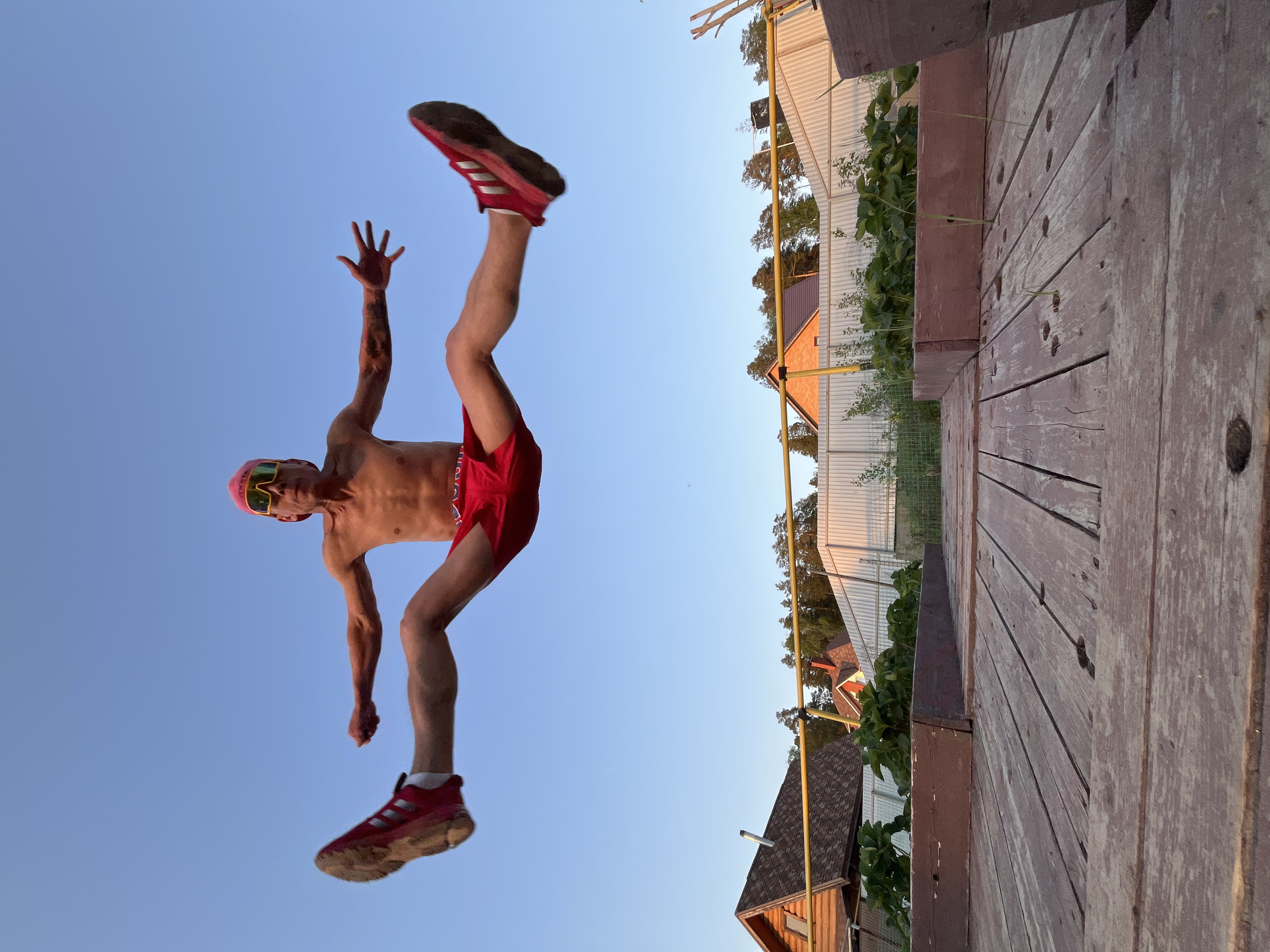 The image captures a man in mid-air against a clear blue sky, seemingly in the city of Chaikovsky, Russia. He is wearing a pink cap, yellow-tinted sunglasses, red shorts, and red sneakers. His arms are outstretched, with one hand open and the other clenched into a fist. The man is shirtless, revealing a slim physique with some visible muscle definition and a tattoo on his right arm. The background includes a wooden deck or platform on the right side of the frame, alongside a partially visible house, a fence, and some greenery. The angle of the shot suggests the photographer was positioned low to the ground, capturing an upward view of the subject.