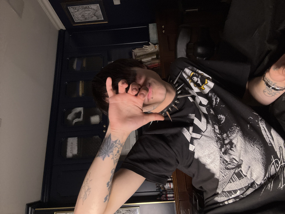 A person with dark hair covers their face with their hand. They wear a black t-shirt with a white graphic design. A spiked collar adorns their neck. The person has several tattoos on their arms and wrist. They sit in what appears to be a dark room with furniture and decorations in the background. The photo is taken from an elevated angle.