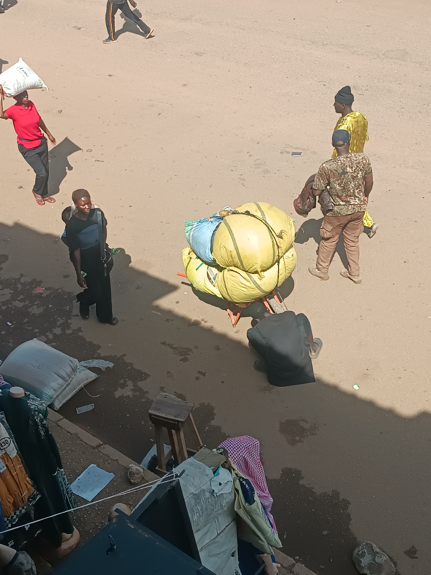 This overhead image depicts a busy outdoor street or market scene under bright daylight. The ground consists of compacted earth and unpaved areas, with prominent shadows indicating strong sunlight.
Several people are engaged in daily activities. In the upper left, a person in a red shirt and dark pants walks to the left, balancing a large white sack on their head. Further down on the left, another person, dressed in dark attire, walks carrying a baby on their back in a sling.
In the center, a handcart is heavily loaded with multiple large yellow and blue sacks. A person in a dark shirt is bent over near the cart, possibly pushing it or adjusting its contents. To the right of the cart, two men stand: one in a vibrant yellow garment and a dark cap, and another in a camouflage-patterned t-shirt and brown trousers. Another individual walks in the background across the upper middle of the frame.
The lower left shows a glimpse of what appears to be a market stall or shop, with clothing items displayed on hangers or mannequins, and a large grey sack resting on the ground. A wooden stool is also visible. Some faint text "430" is discernible on a clothing tag. The scene portrays typical commerce and movement of goods in a lively outdoor environment.