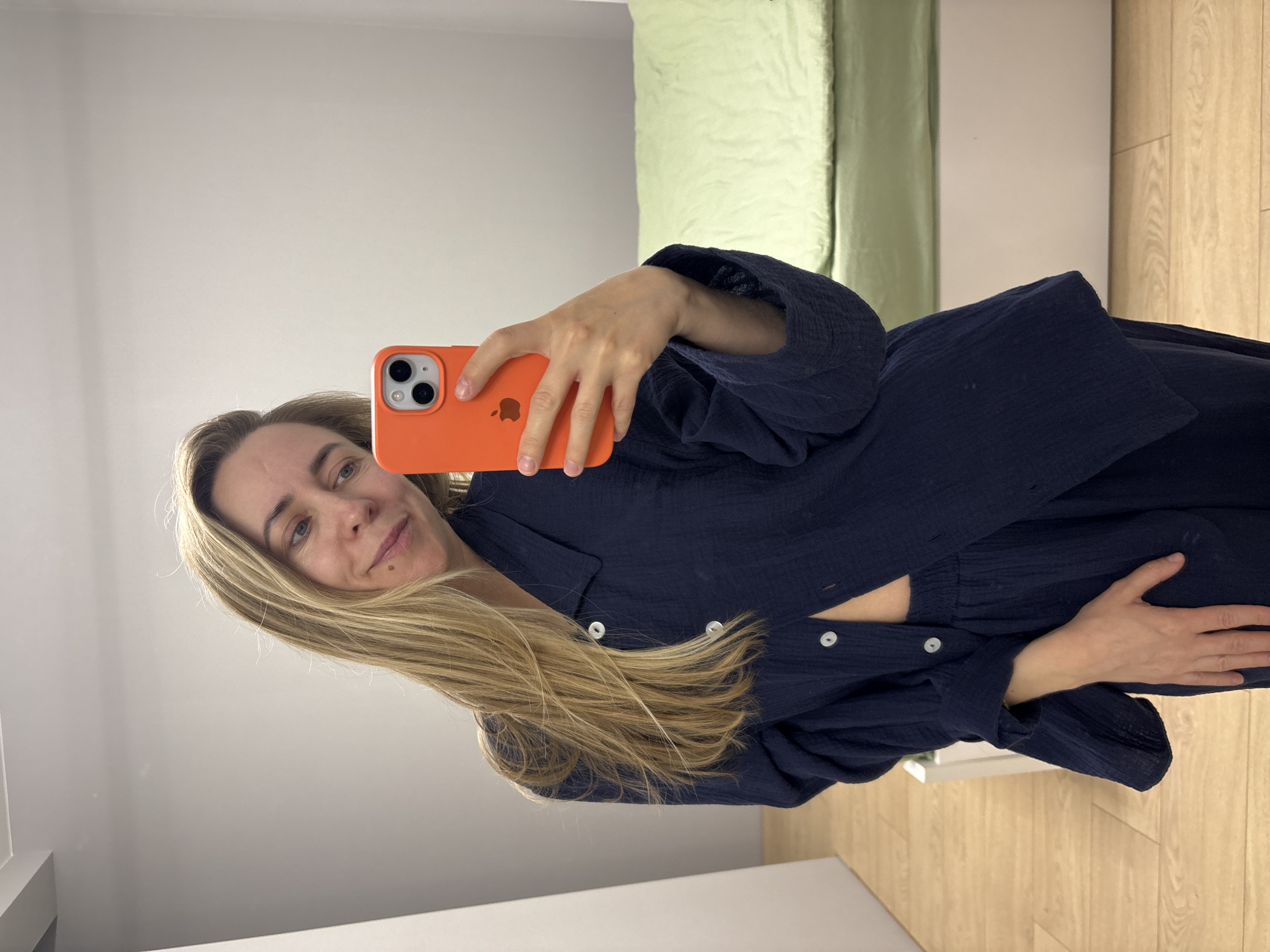 The image captures a woman taking a mirror selfie indoors, likely in a bedroom or dressing area in Chaikovsky, Russia. The woman, with long, light blonde hair that falls over her shoulders, is visible from the chest up, angled slightly to her left. She holds a bright orange-cased smartphone, appearing to be an iPhone (indicated by the camera layout and a visible Apple logo on the case), with her right hand to capture her reflection. She has a subtle, pleasant smile and a small mole visible on her left cheek.
She is wearing a dark blue, possibly navy, long-sleeved button-up top made of a textured fabric, possibly gauze or crinkle cotton, featuring several prominent white buttons down the front. The top appears to be loose-fitting and casual.
The background, reflected in the mirror, shows a clean, modern interior. On the left side, there's a plain, light-colored wall. To the right, a pale green textile, possibly a curtain or bedspread, is visible, alongside a white surface which might be a bed frame or furniture. The lower portion of the reflection reveals light-colored wooden flooring. The lighting in the room appears bright and even, suggesting either natural daylight or good artificial illumination.