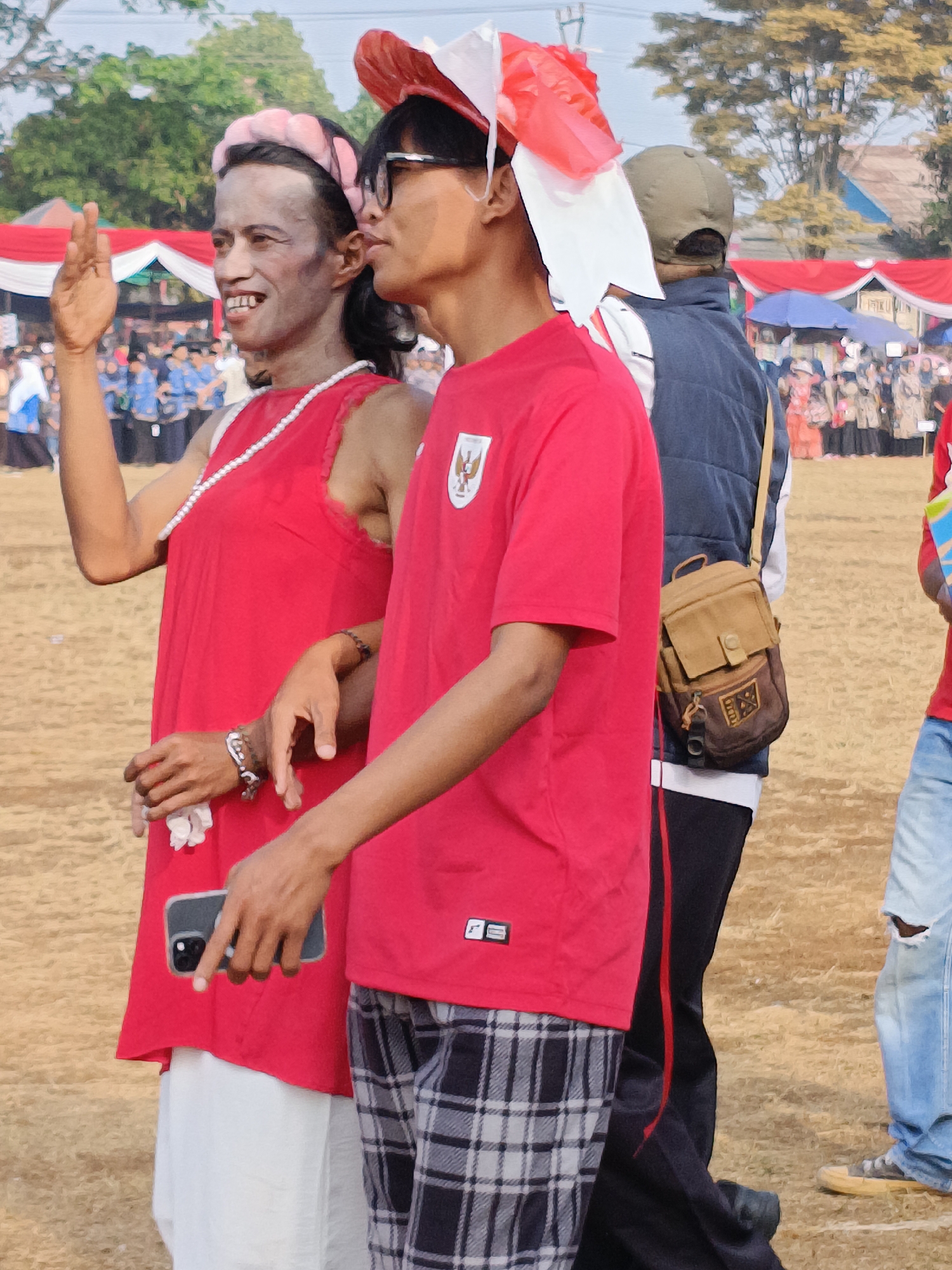 The image captures a street-level scene in Cibodas, Indonesia, focusing on two individuals in the foreground. One person, facing left, wears a red dress with a pearl necklace and a pink headband. They are holding a mobile phone in their left hand and have their right hand raised in a wave. Their facial expression is a wide smile. The other individual, to the right, is wearing a red t-shirt with a crest and black and white plaid pants. They are looking to the right, and on their head, they have a red and white festive headpiece.
Behind them, the background suggests an outdoor event. Red and white banners are visible, along with tents and a crowd of people in the distance, some holding umbrellas. Trees and buildings can be seen further in the background under a bright sky. The ground is a dusty, light brown color. The overall atmosphere appears to be celebratory or part of a public gathering.