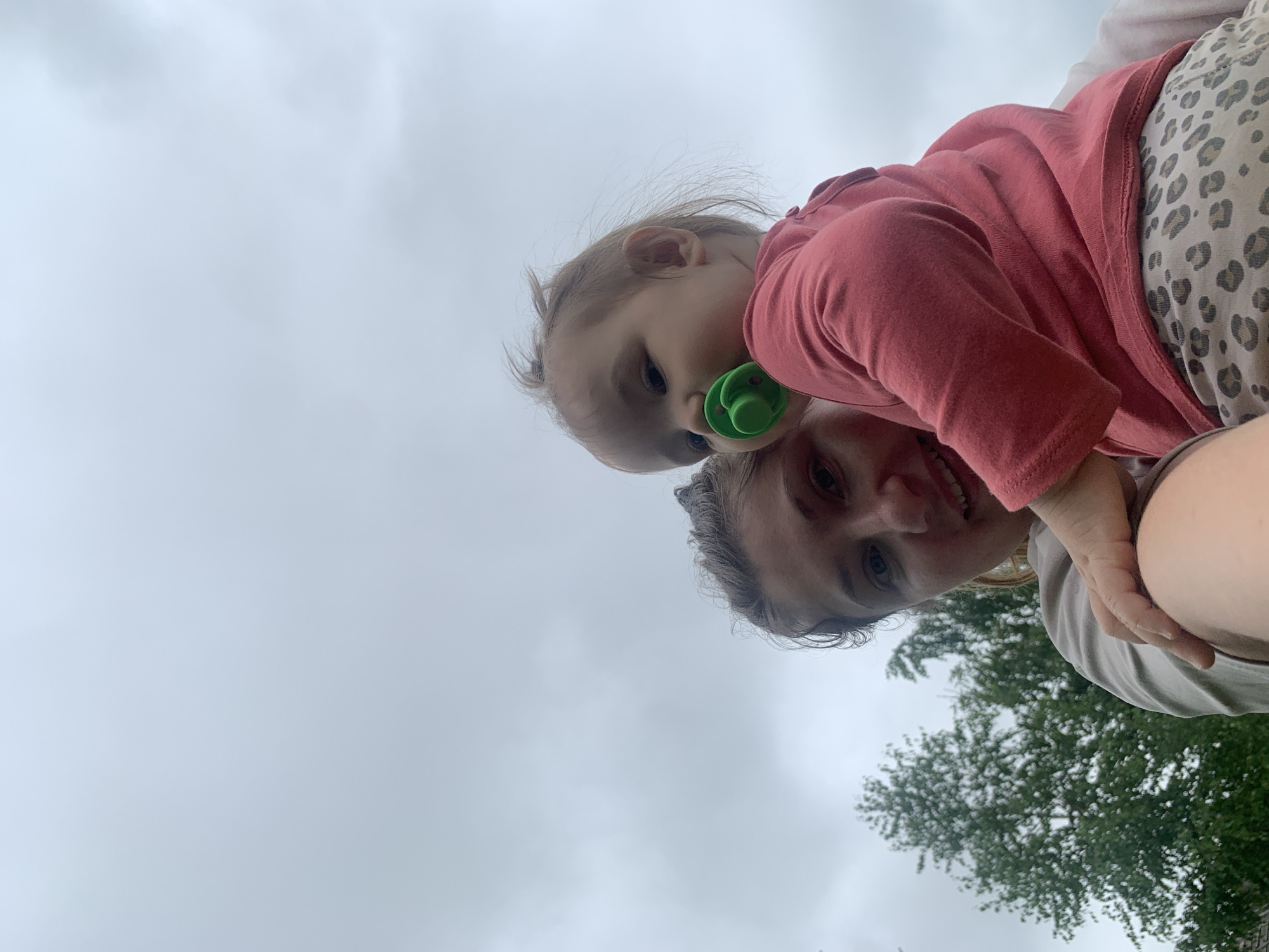The image captures a woman holding a baby against a cloudy sky. The woman is in the foreground on the left, looking towards the camera with a slight smile. She is wearing a light-colored t-shirt. The baby is in her arms, facing away from the camera and to the right, with a green pacifier in its mouth. The baby is wearing a pink long-sleeved shirt and leopard-print bottoms. In the background, trees are visible on the left side of the image. The setting appears to be outdoors, likely a residential area, with no specific identifying landmarks of Zasechnyi, Russia.