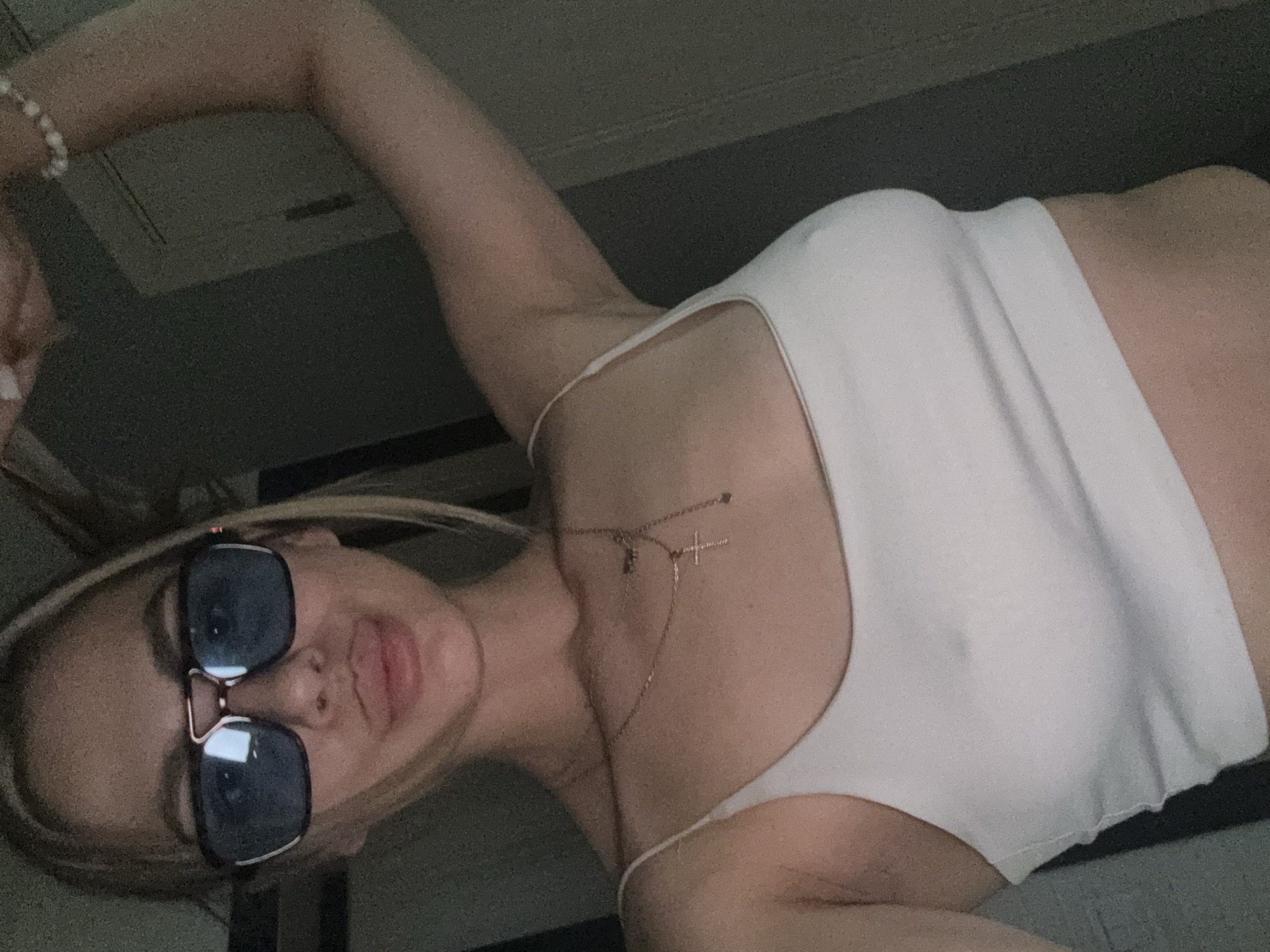 The media file is a close-up, rotated selfie of a young woman. She is wearing a white tank top and large, dark-lensed sunglasses. A delicate gold necklace with a small cross pendant rests on her chest. Her hair is blonde and appears to be pulled back. The background is dark and out of focus, with a hint of what might be a ceiling or wall. The lighting suggests it could be indoors, possibly at night or in a dimly lit room. The woman's expression is casual, with her mouth slightly open. The image appears to have been taken at a low angle. There is no visible text in the image, and no specific information about the activity or event taking place is discernible.